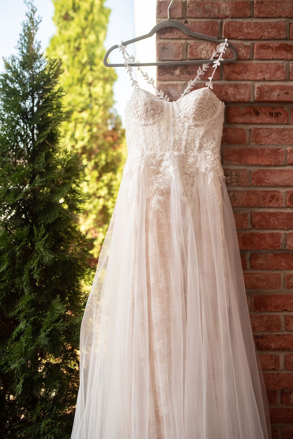 Strapless lace and tulle blush wedding gown hanging on a hanger against a rustic brick wall outdoors.