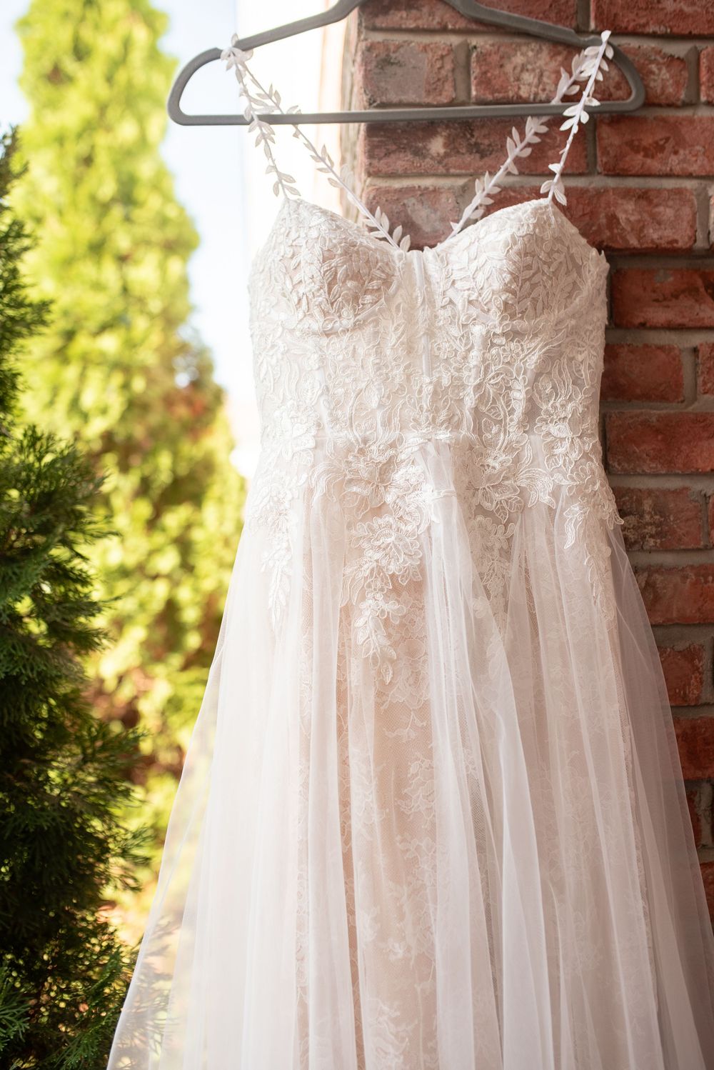 Elegant white lace wedding dress with spaghetti straps hanging on a hanger against a brick wall.