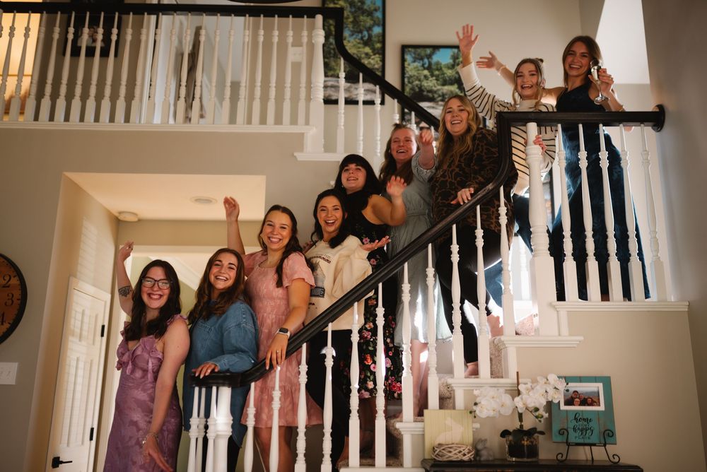 A joyful group of women in colorful dresses celebrating on a staircase, waving and smiling at a wedding party.