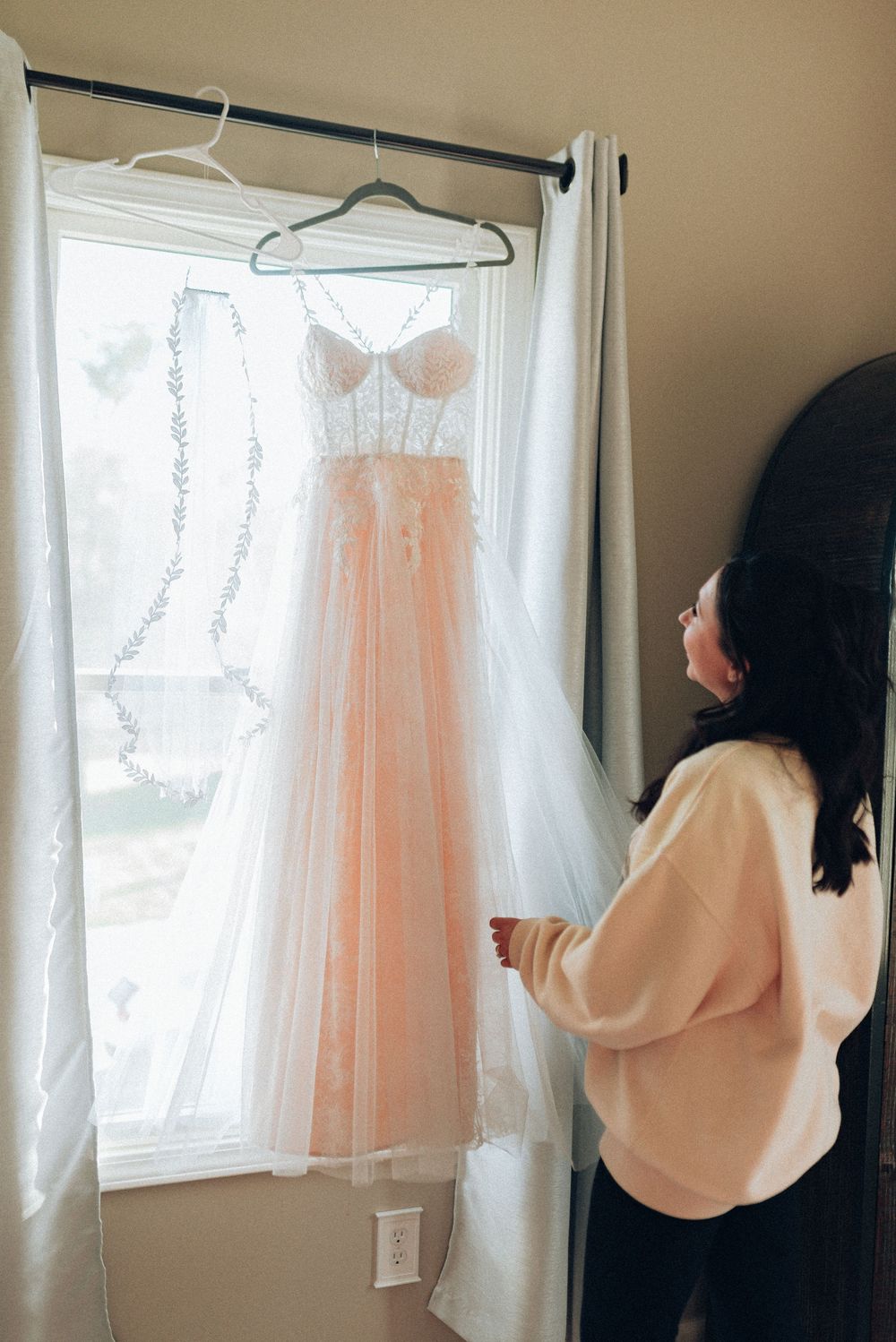 Bride admiring her blush lace and tulle wedding dress hanging in a sunlit window before the ceremony.