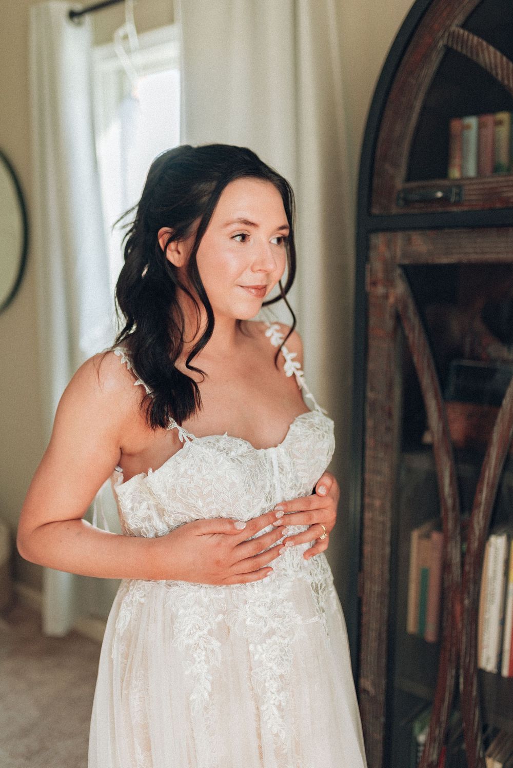 Brunette bride in lace wedding gown adjusting dress near rustic wooden bookshelf.