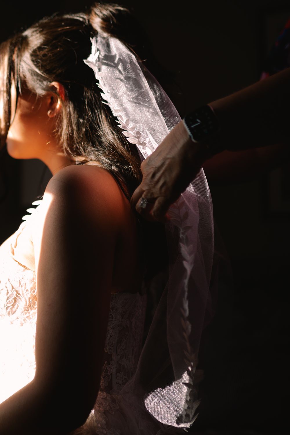 A bride in a lace wedding dress having her veil adjusted by a gloved hand in dramatic lighting.