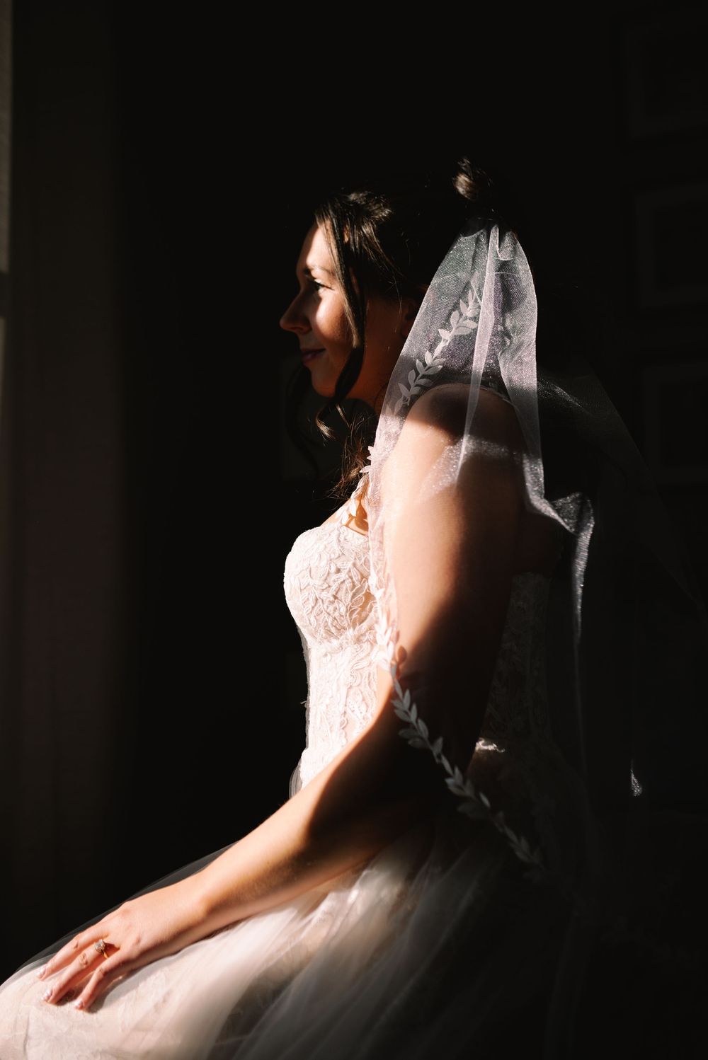 Bride in a white lace wedding dress and veil sitting in dramatic window light, profile view.