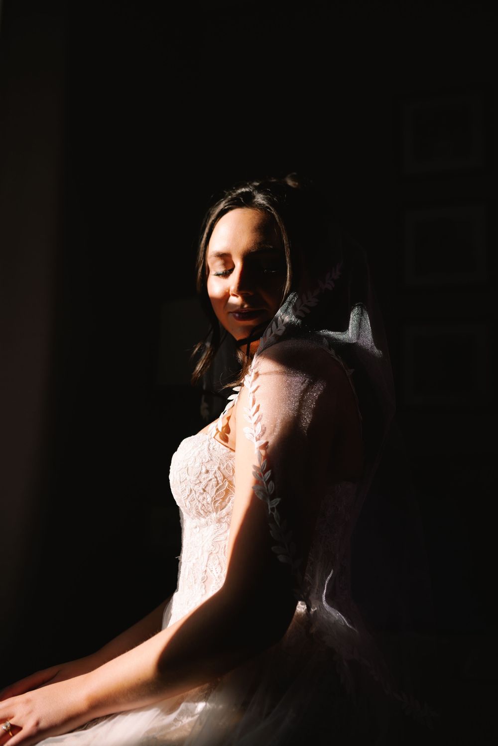 Beautiful bride in a lace wedding gown sitting peacefully with eyes closed in dramatic moody lighting.