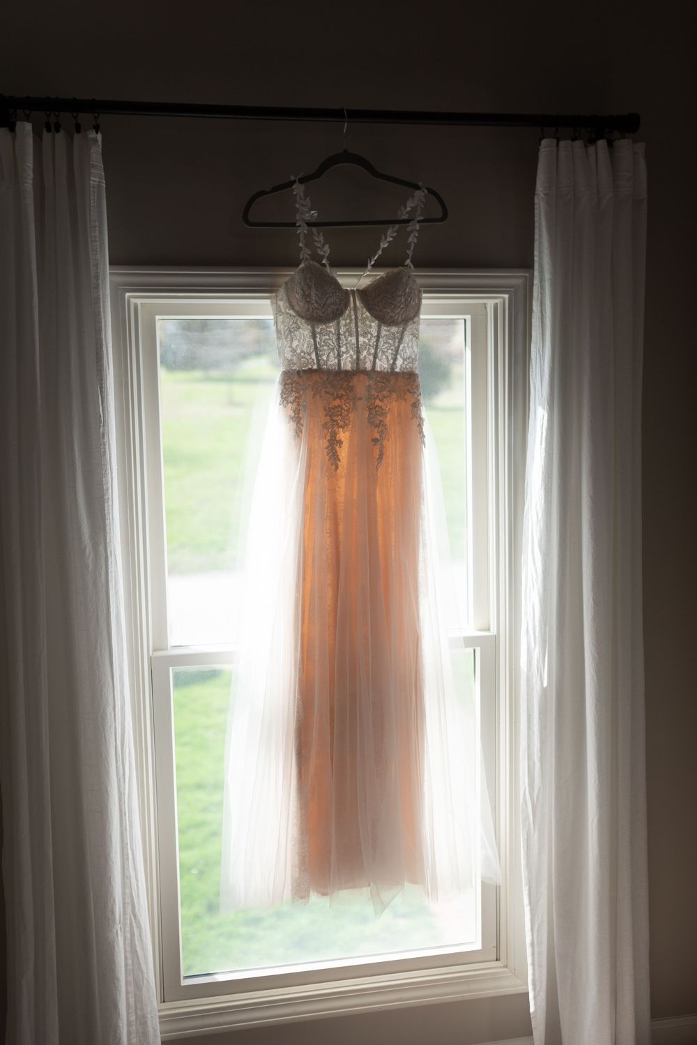 Elegant blush wedding dress with lace bodice hanging on a curtained window, backlit by natural light.