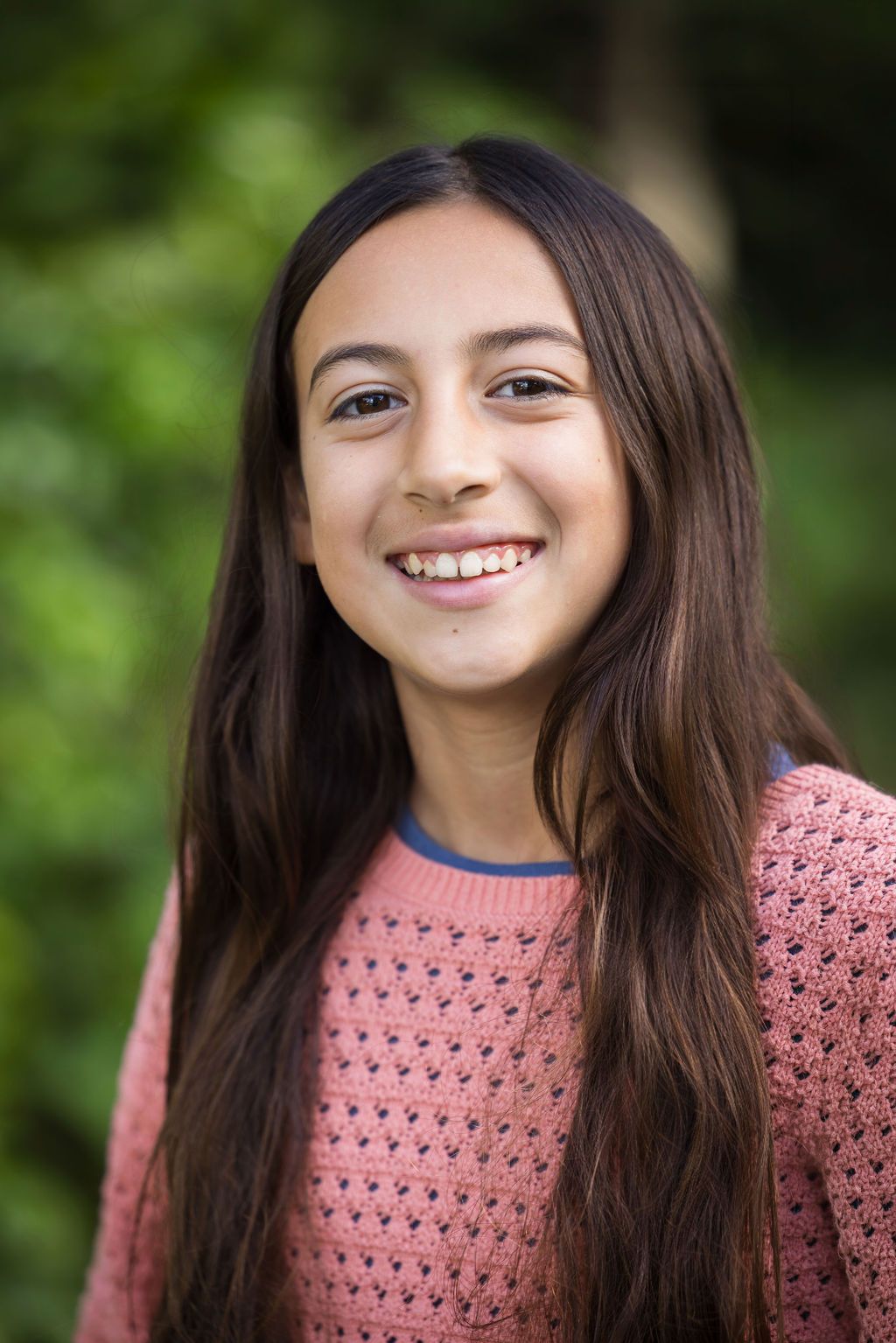 Smiling young girl with long brown hair wearing a pink sweater outdoors.