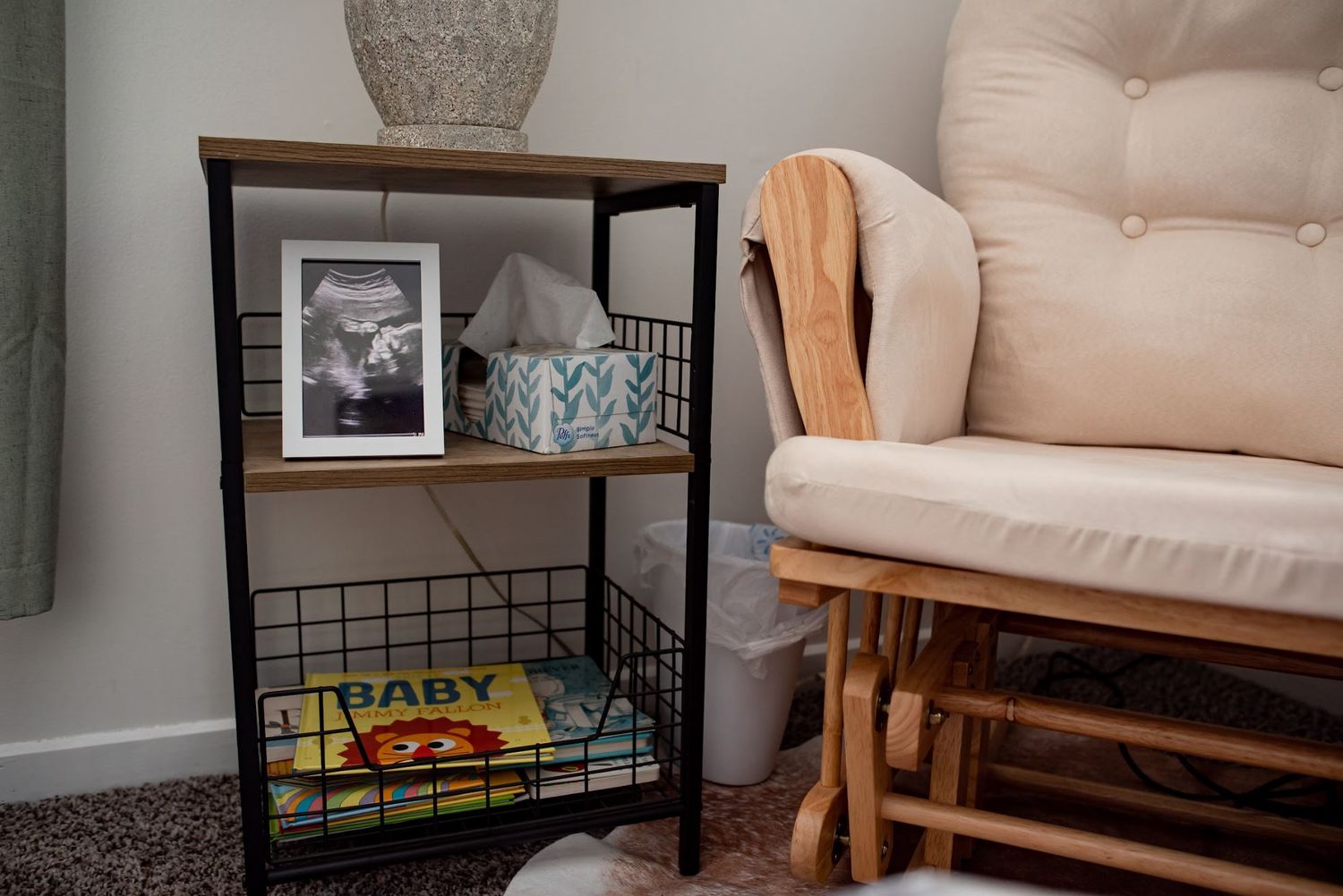 Cozy nursery corner with beige chair, framed photos, and wooden storage shelf.