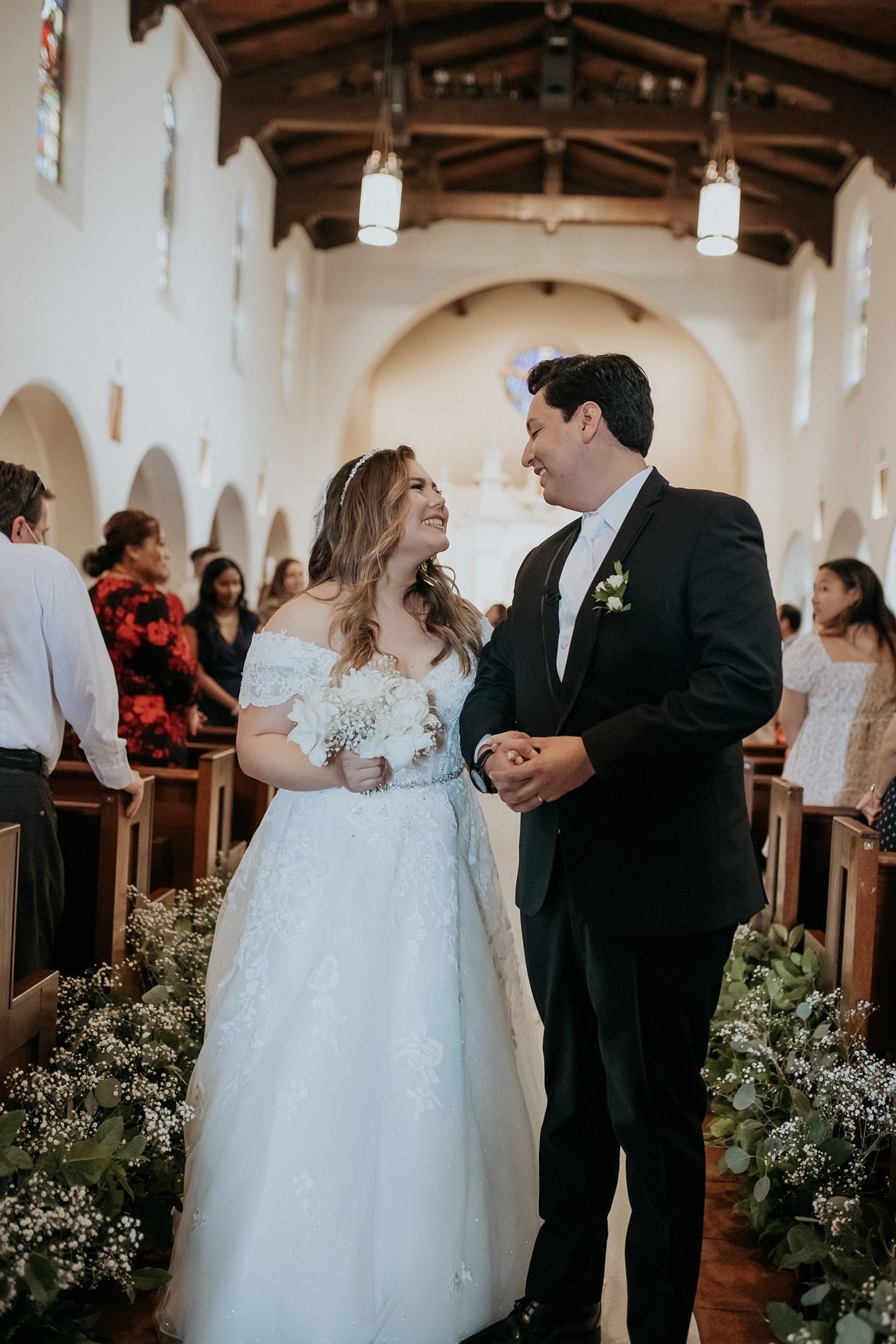Bride and groom smiling at each other while walking down the aisle of a white church decorated with floral arrangements.