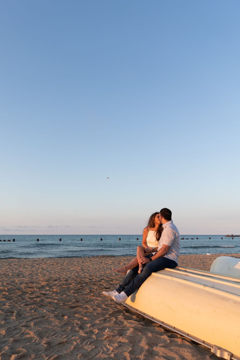 Couple sharing a romantic kiss while sitting on a boat on a sandy Chicago North Avenue beach at sunset with calm lake Michigan behind them.