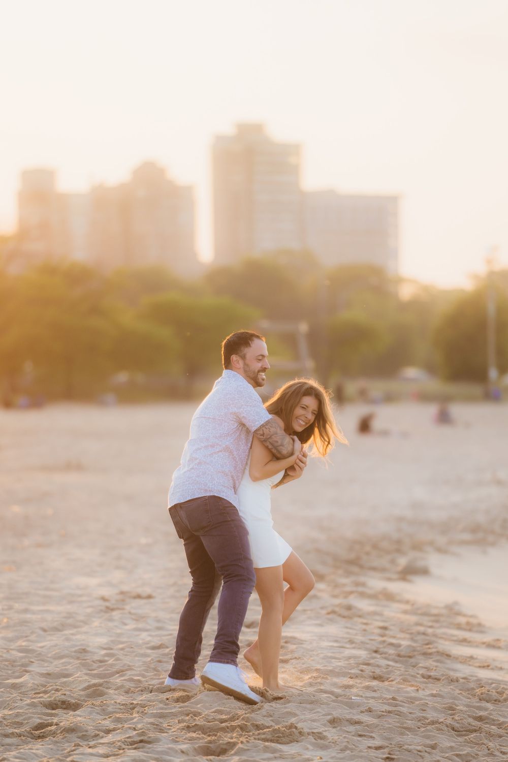 Couple laughing and embracing playfully on a sandy North Avenue beach at golden hour with Chicago skyline in background.