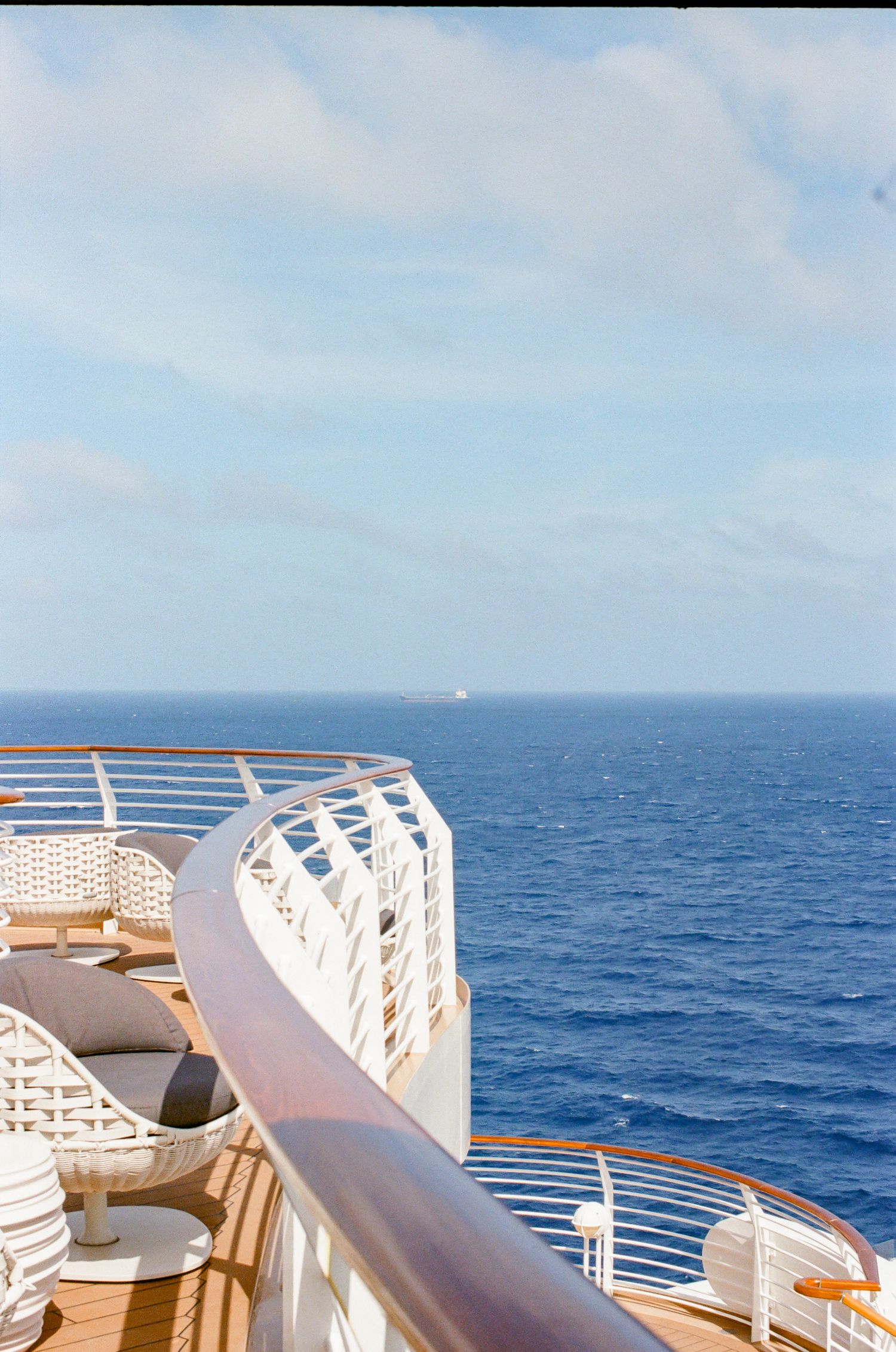 Luxury cruise ship deck with white railings, wooden floors, and lounge chairs overlooking the vast blue ocean horizon.