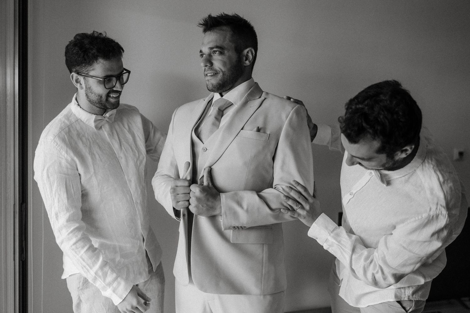 Groom in light suit being helped by two groomsmen while getting ready for wedding ceremony.