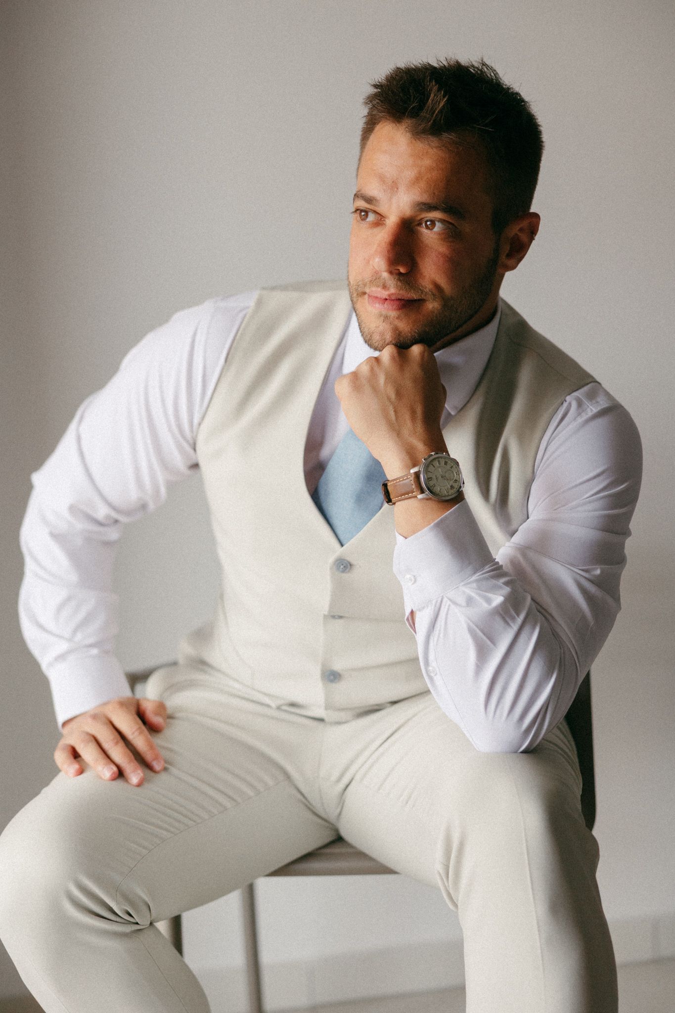A well-dressed man in a beige vest and white shirt sits confidently, resting his chin on his hand, wearing a watch.