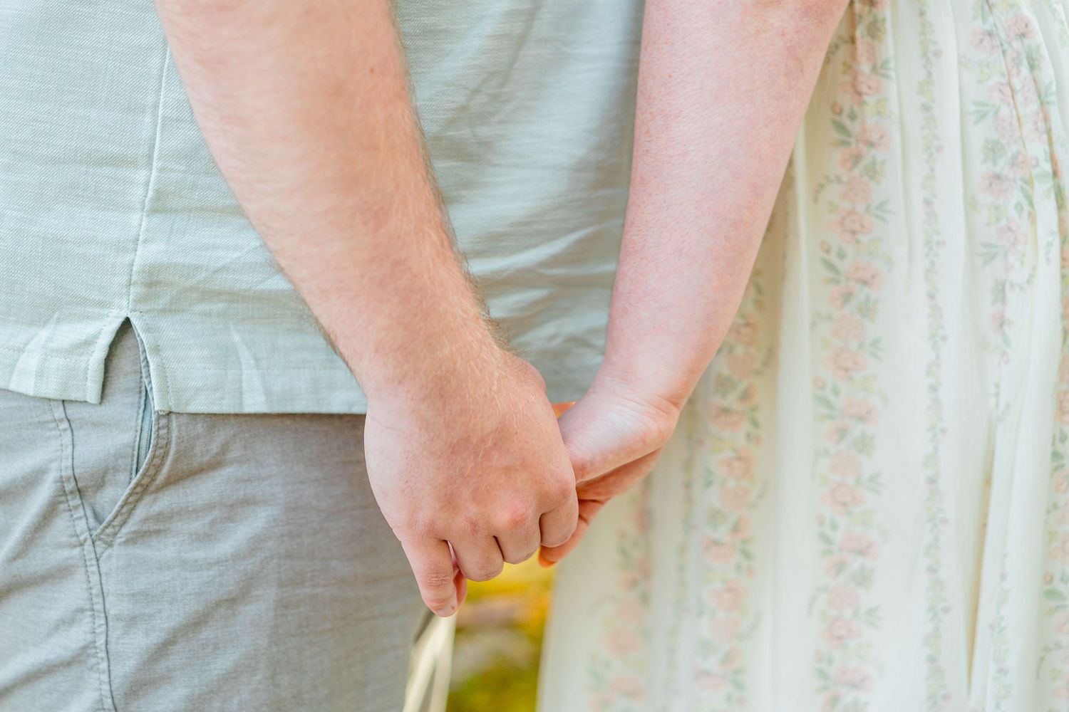 Couple holding hands outdoors, man in gray shirt and pants, woman in floral dress, romantic close-up shot.