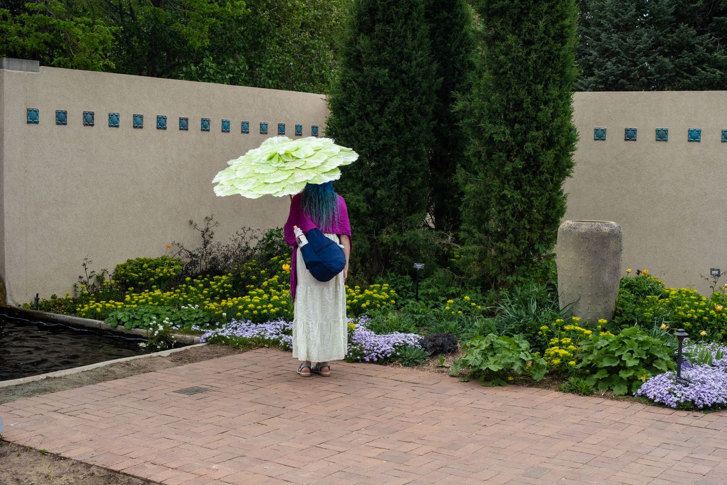 Woman holding a decorative green lotus umbrella in a colorful garden with cypress trees and adobe wall.