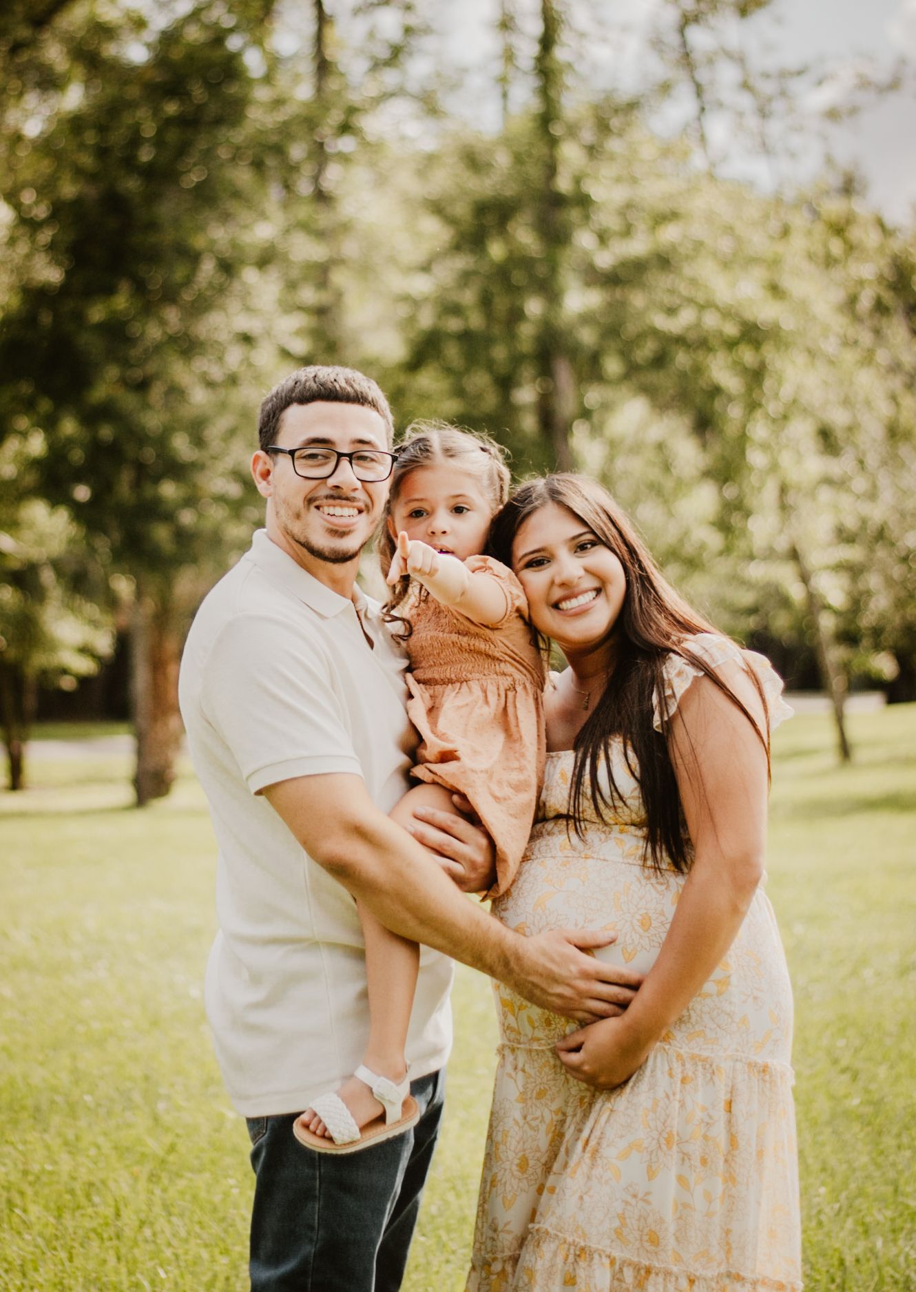 A smiling couple holding their toddler daughter outdoors in a green park, mother visibly pregnant.