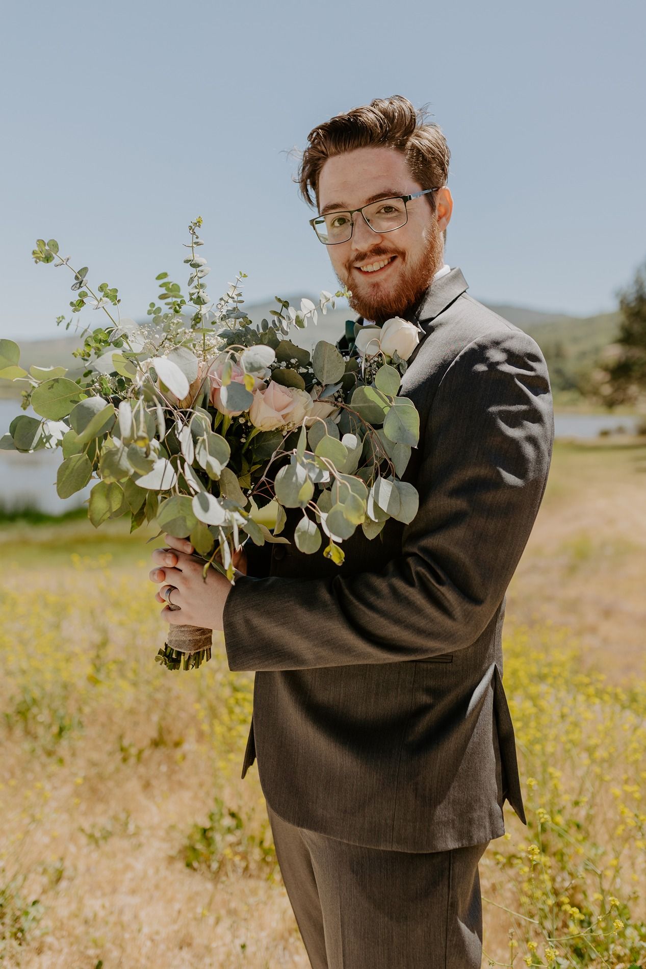 Smiling groom in gray suit holding large floral bouquet with eucalyptus and roses outdoors on sunny day at Lake Cuyamaca, Julian.