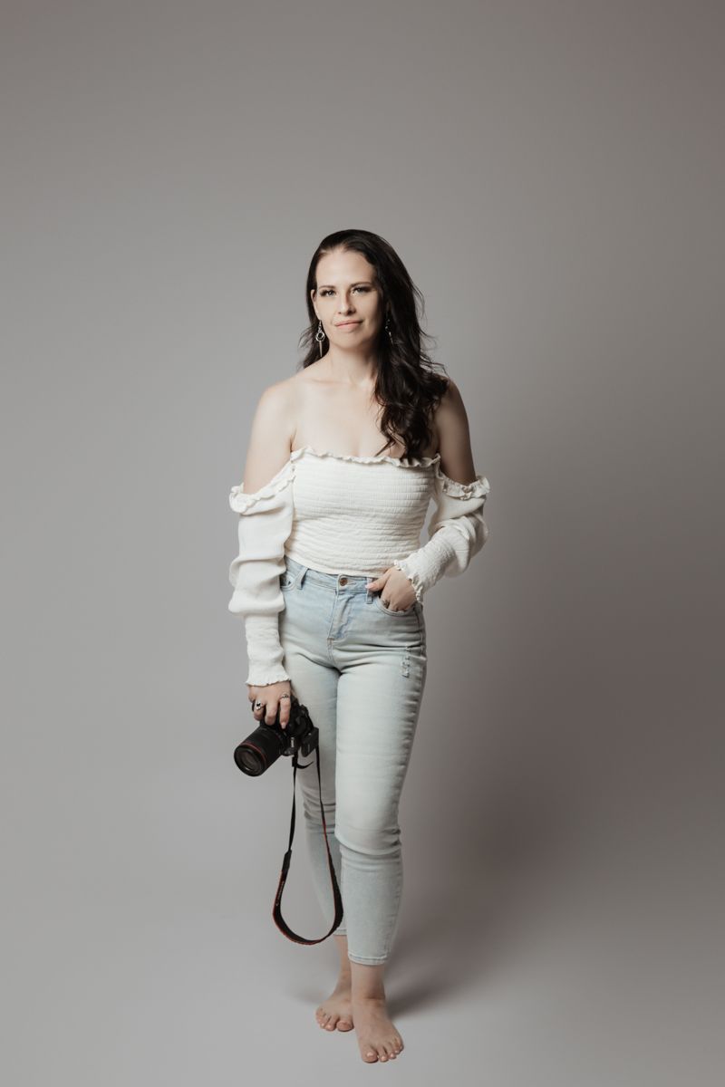 Female wedding photographer standing barefoot holding camera in white off-shoulder top and jeans.