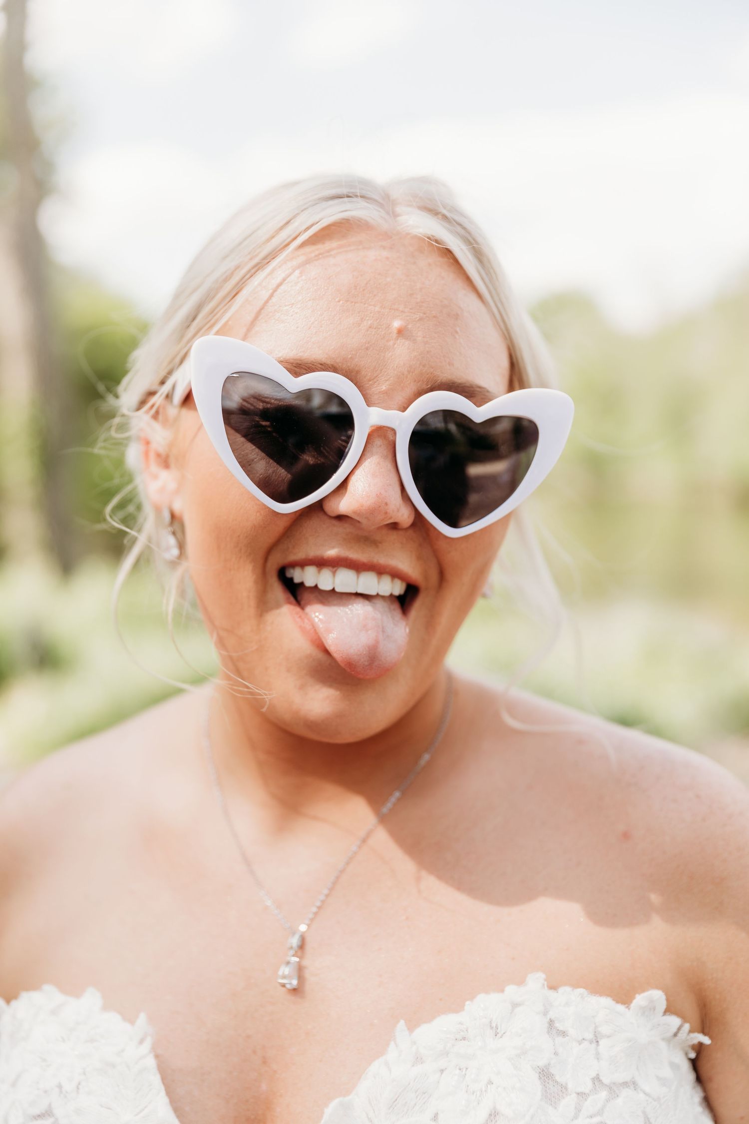 Bride in white lace dress wearing heart-shaped sunglasses sticking out tongue, smiling outdoors.