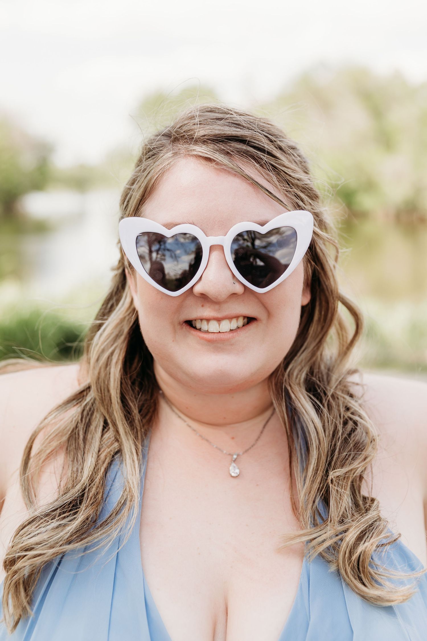 Smiling woman wearing white heart-shaped sunglasses and blue dress with a pendant necklace outdoors.