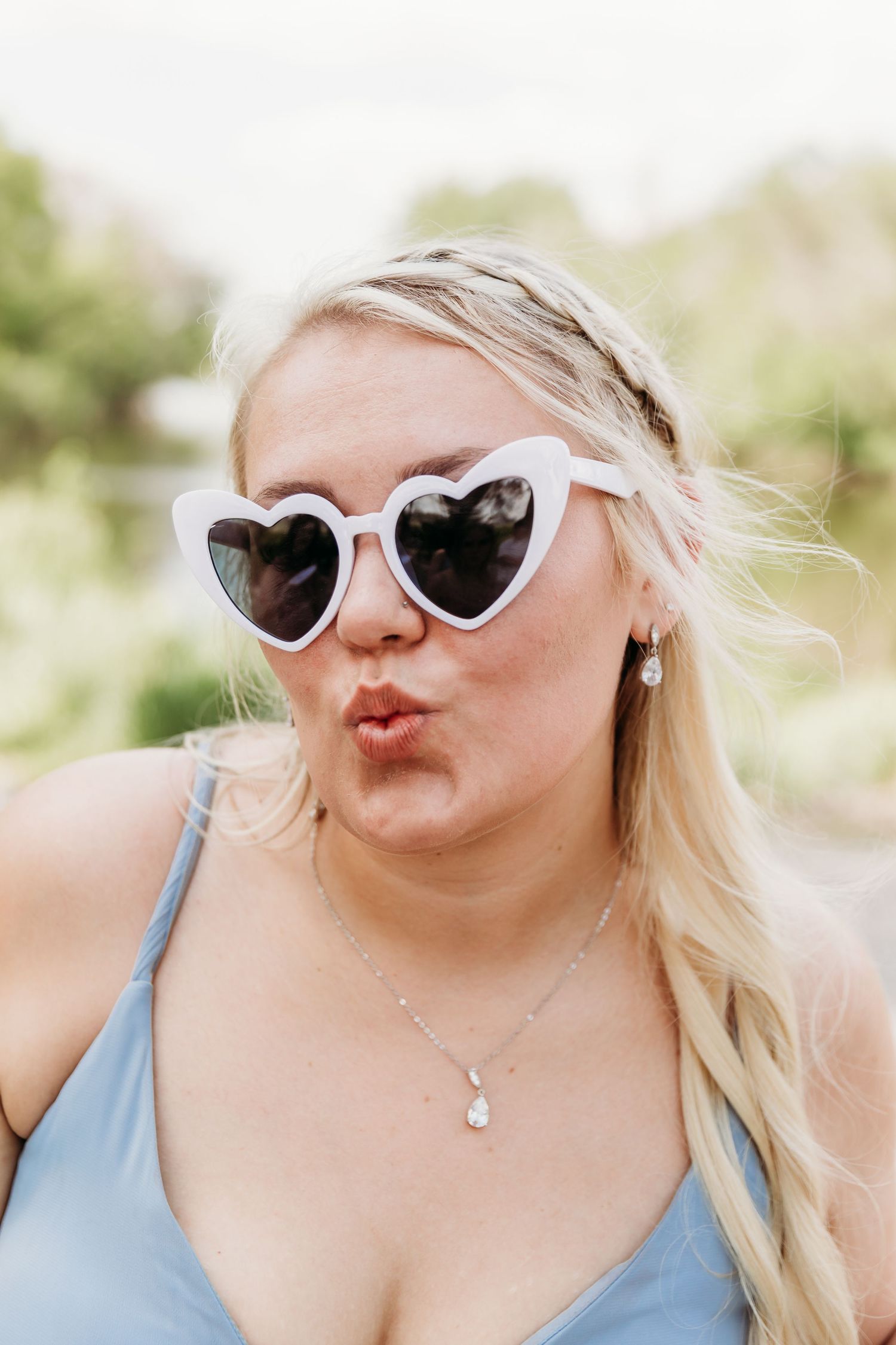 Blonde woman wearing white heart-shaped sunglasses making a kissing face, wearing a blue top and silver necklace.