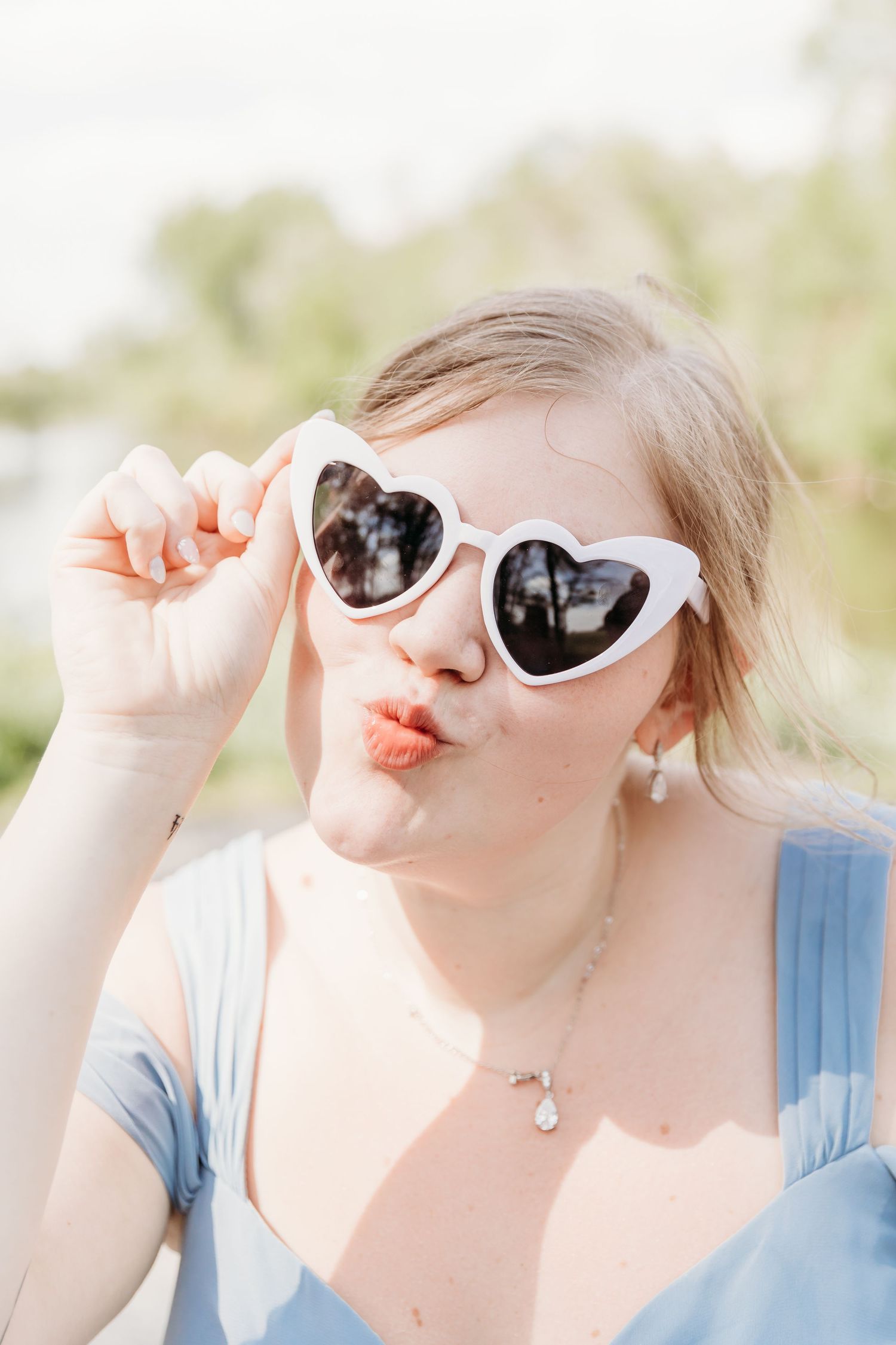 Young woman in heart-shaped white sunglasses making a kissing face outdoors in a light blue dress.