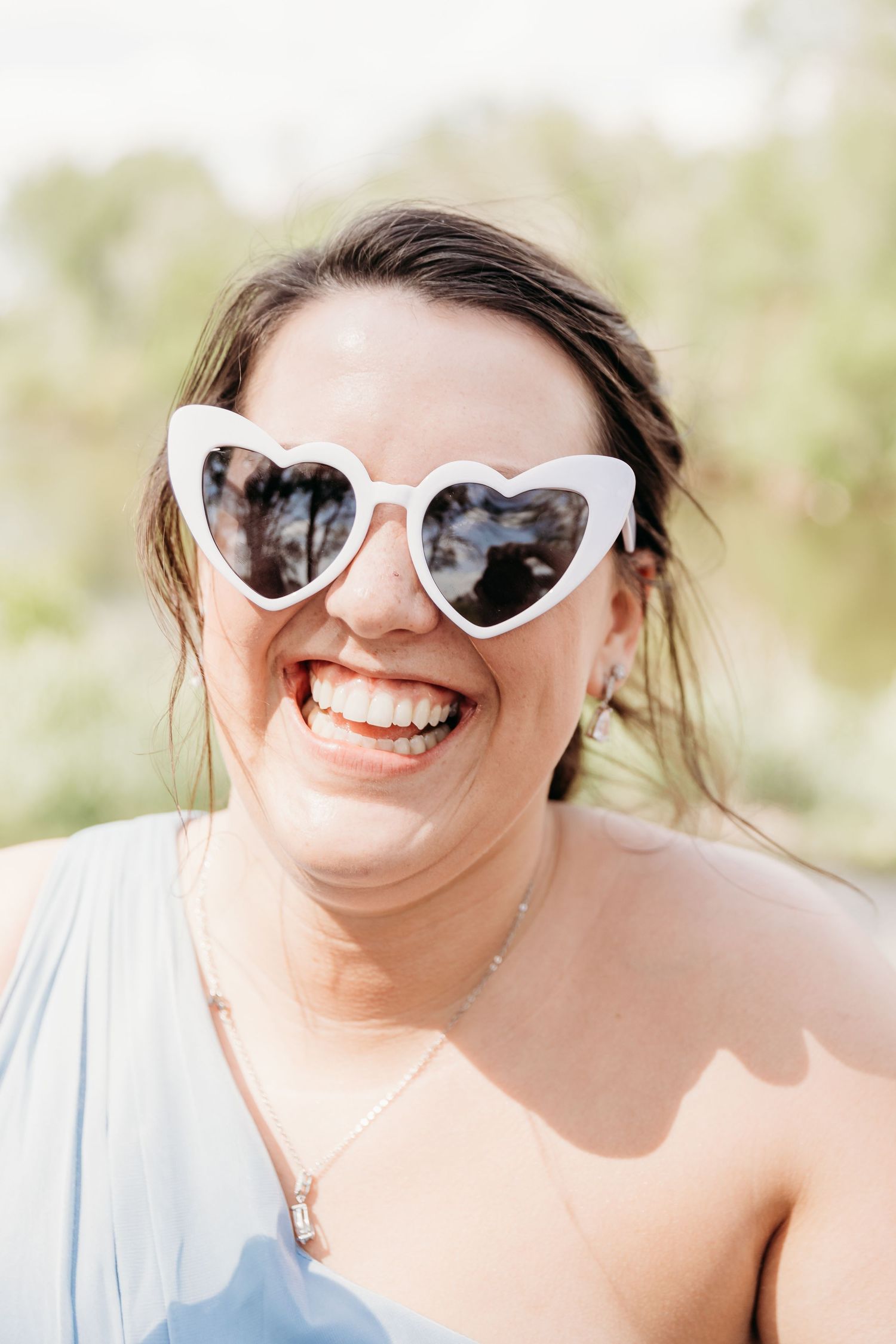 A woman laughing joyfully wearing white heart-shaped sunglasses and a light blue dress outdoors.