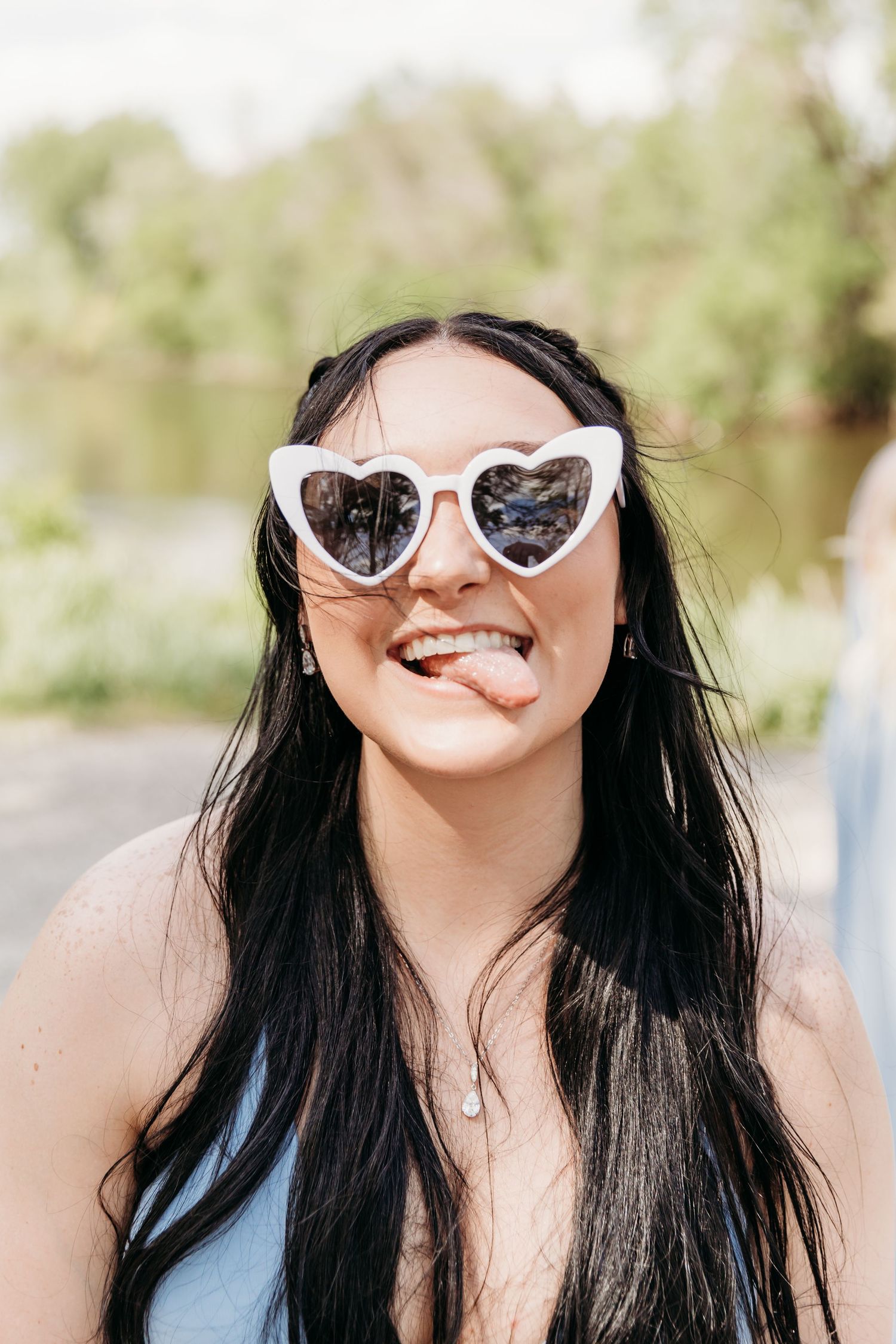 Young woman with long dark hair wearing white heart-shaped sunglasses sticking out tongue outdoors.