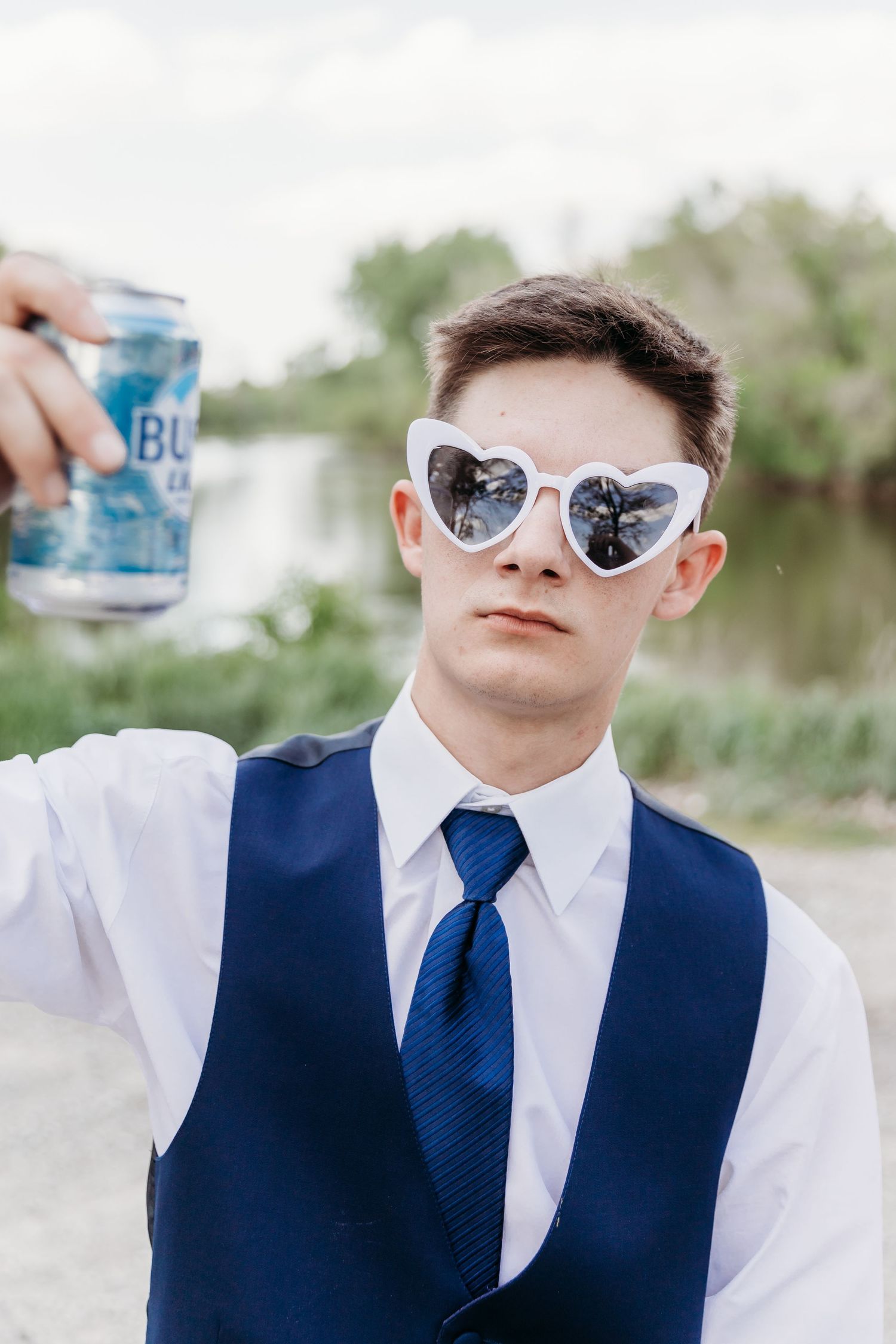 Young man in blue vest and tie wearing heart sunglasses holding up a can outdoors.