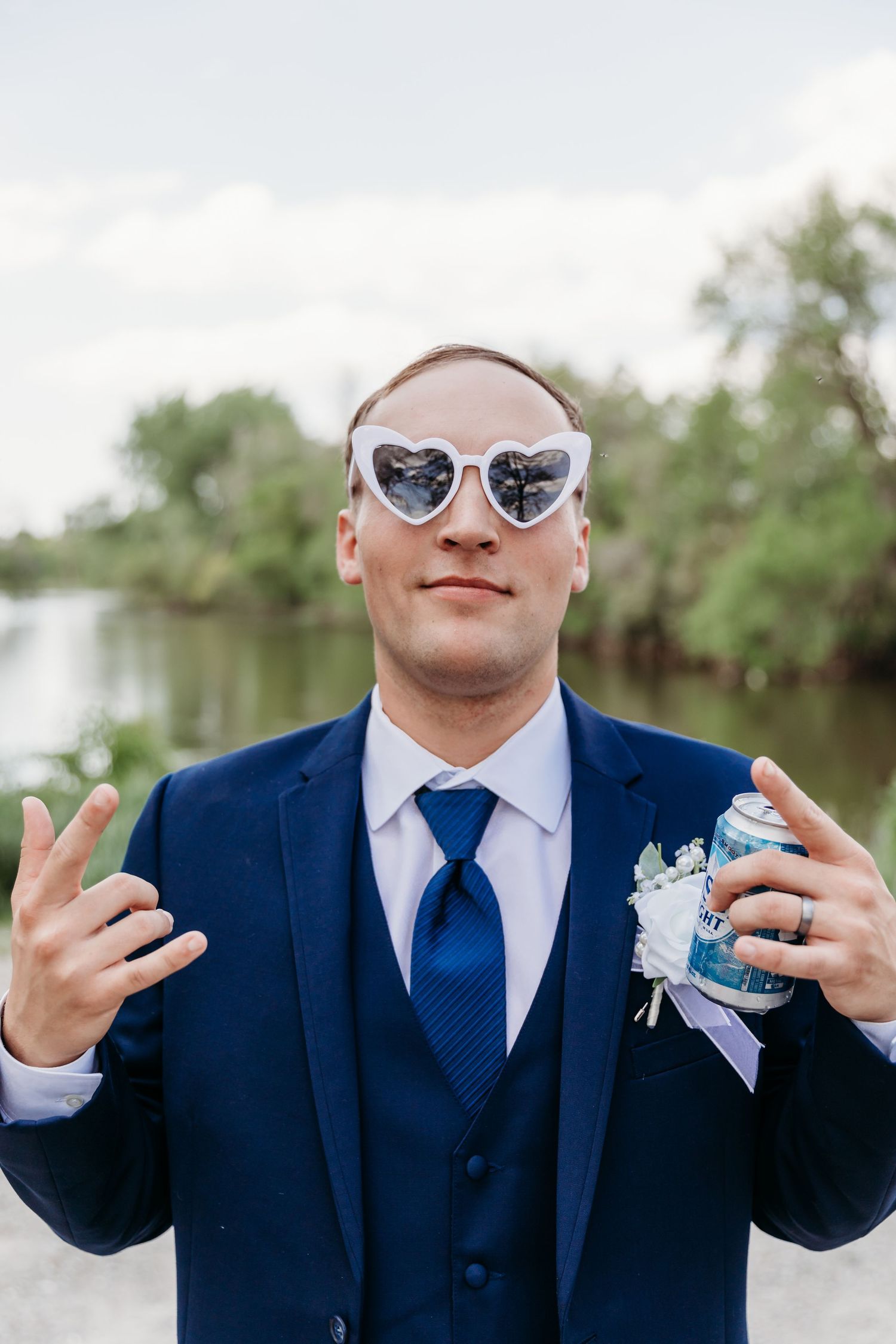 Groom in navy suit and heart-shaped sunglasses holds a drink, flashing peace signs outdoors near a river.