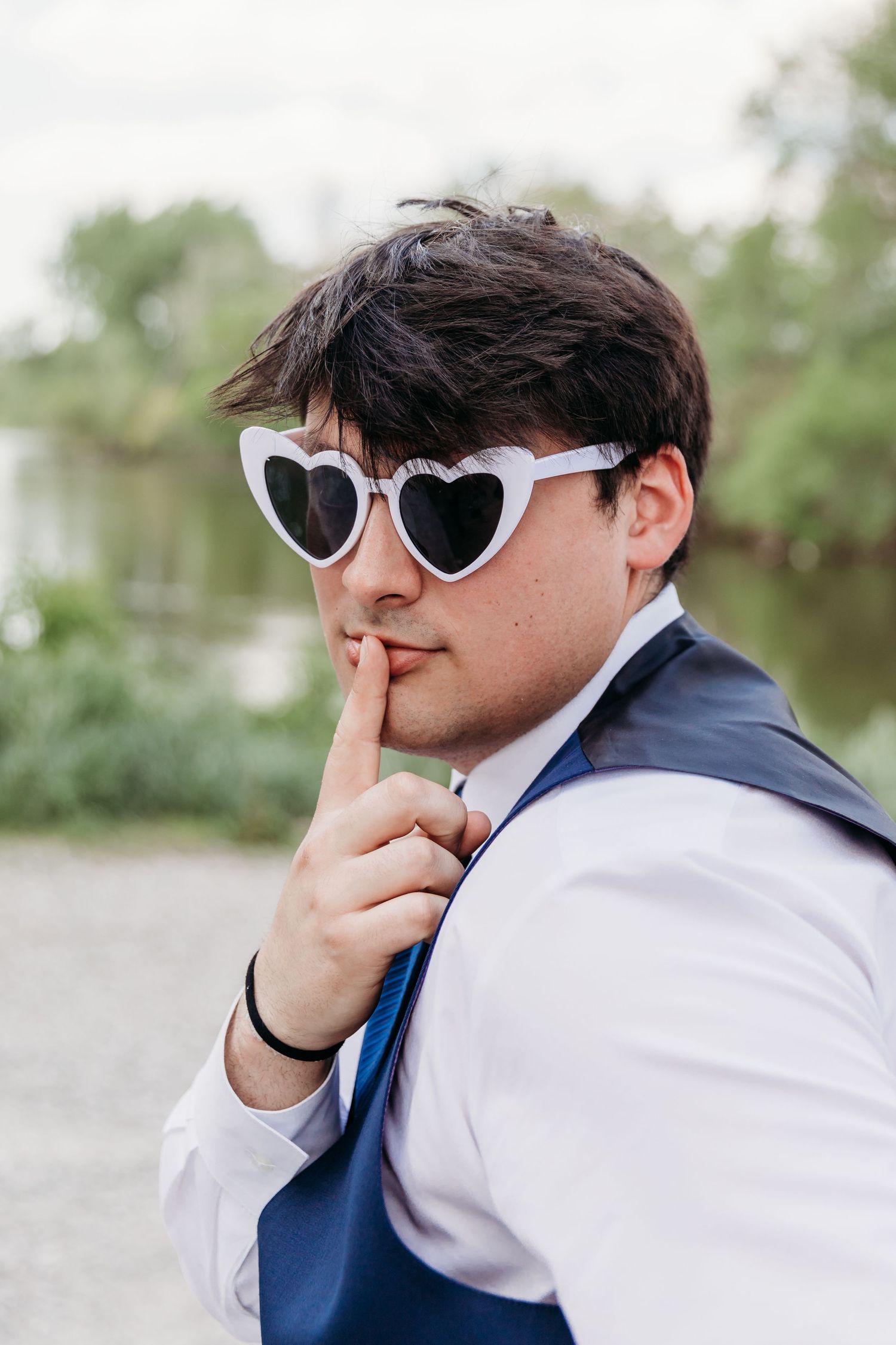 Young man in white heart sunglasses, blue vest and tie, making a shush gesture outdoors.