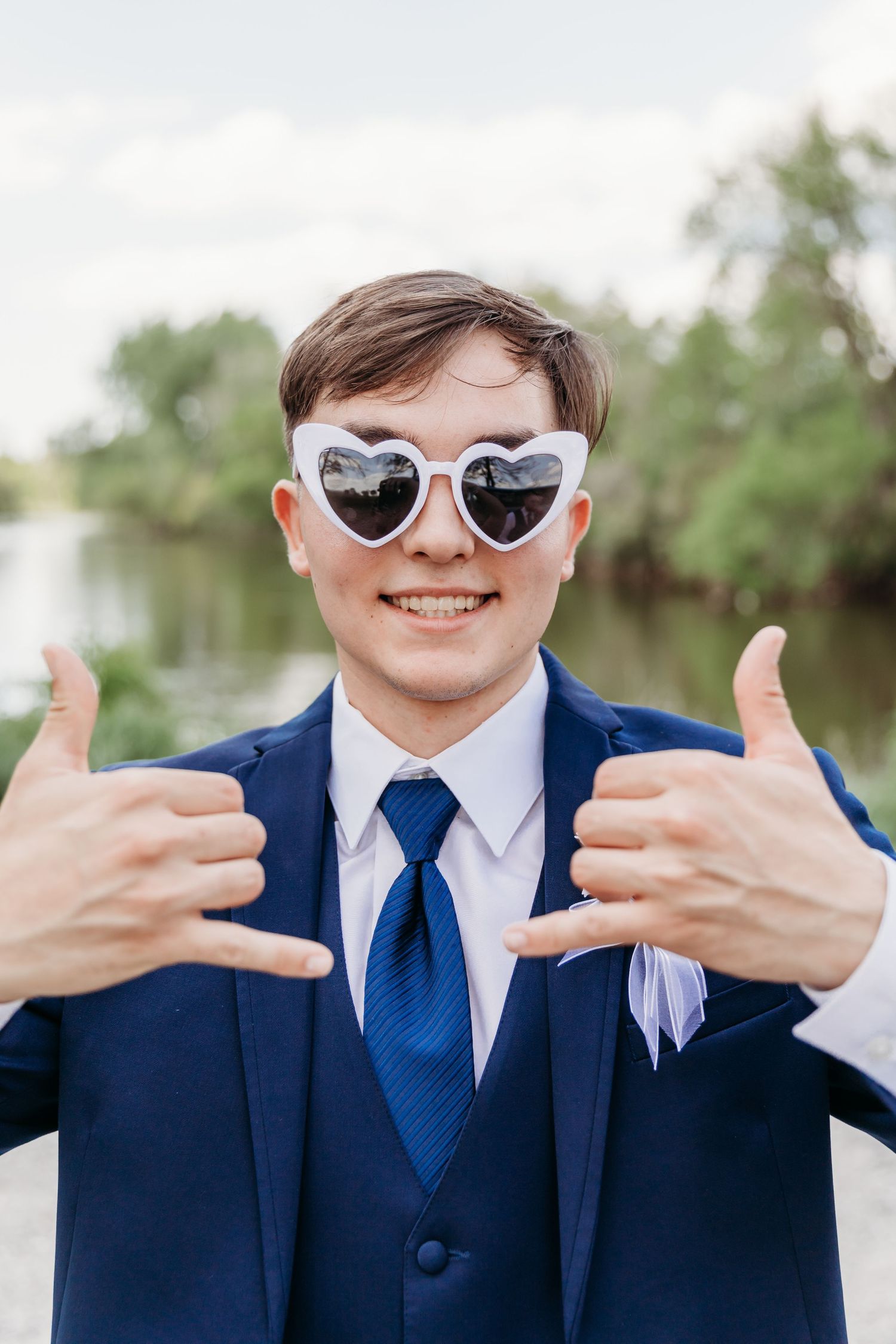 Young man in navy suit and heart-shaped sunglasses giving thumbs up outdoors.