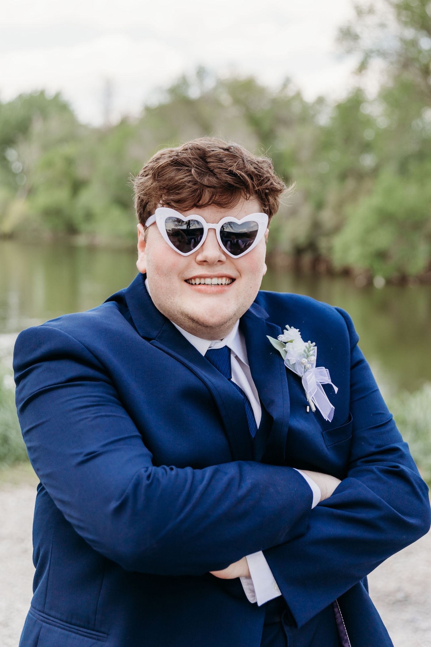 Smiling young man in navy suit with boutonniere and heart-shaped sunglasses, arms crossed.