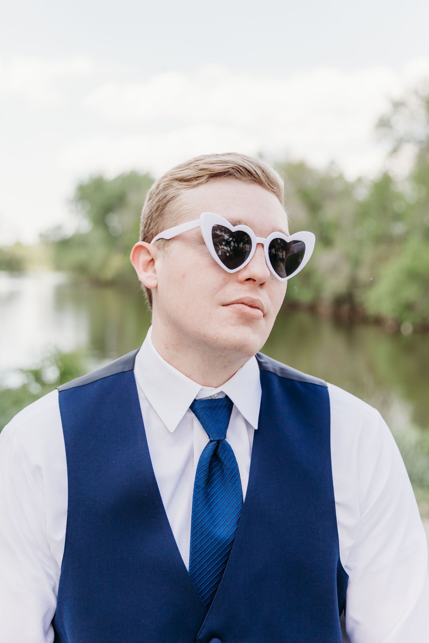 Young man wearing white heart-shaped sunglasses, blue vest and tie, posing outdoors near water.