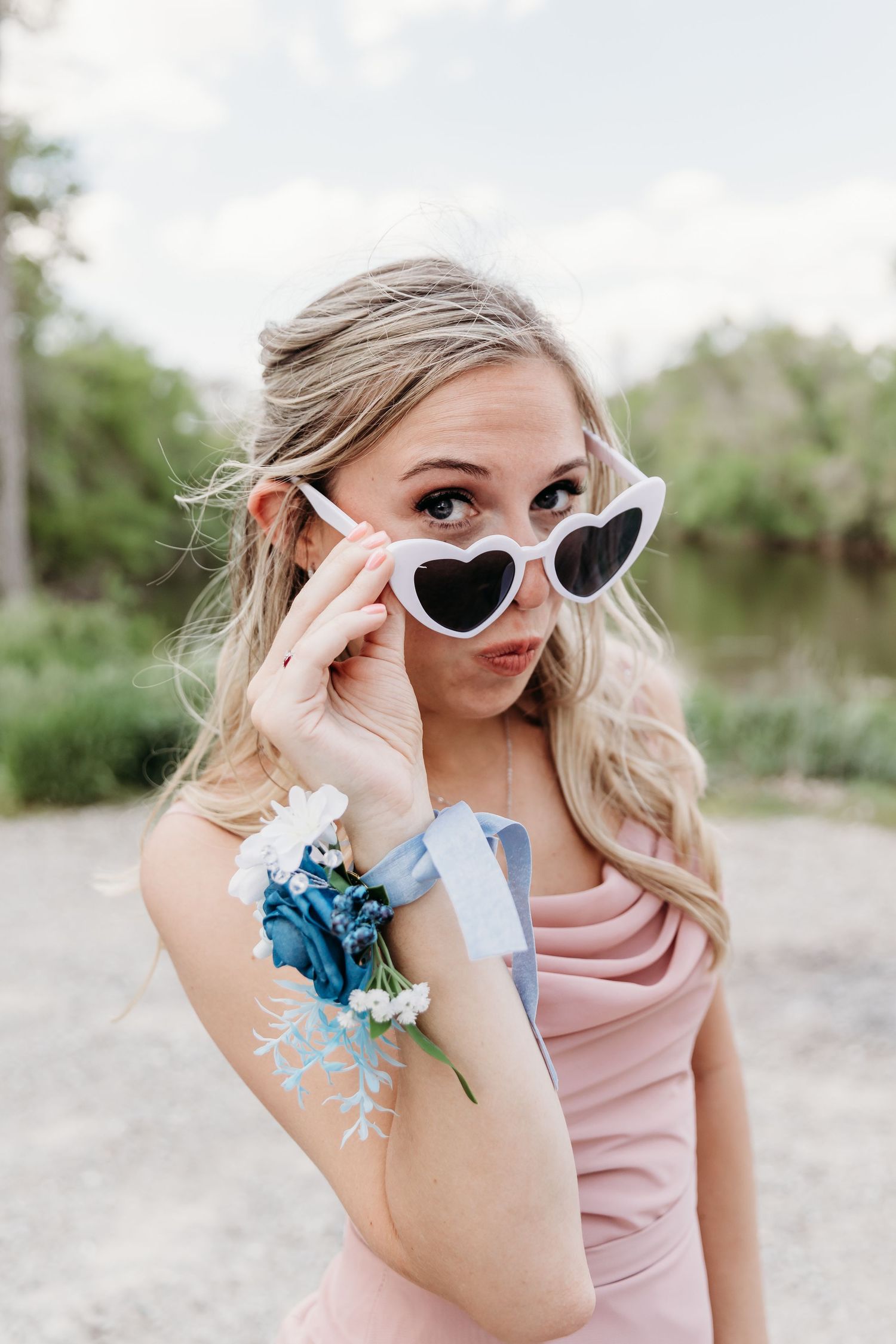 Young woman in pink dress wearing heart-shaped sunglasses with a floral wrist corsage, posing outdoors.