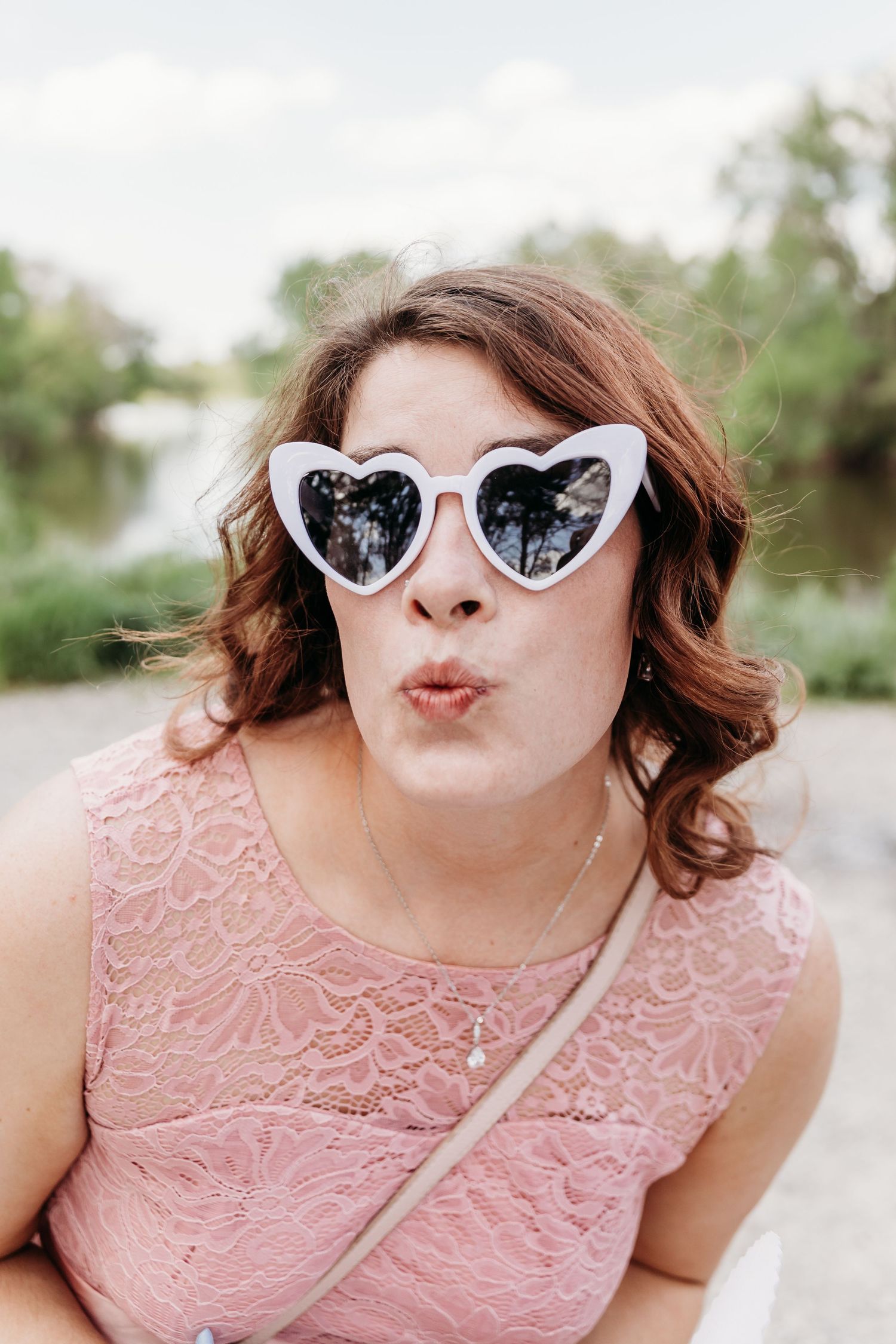 Woman with curly brown hair in pink lace top wearing white heart sunglasses making kissy face.