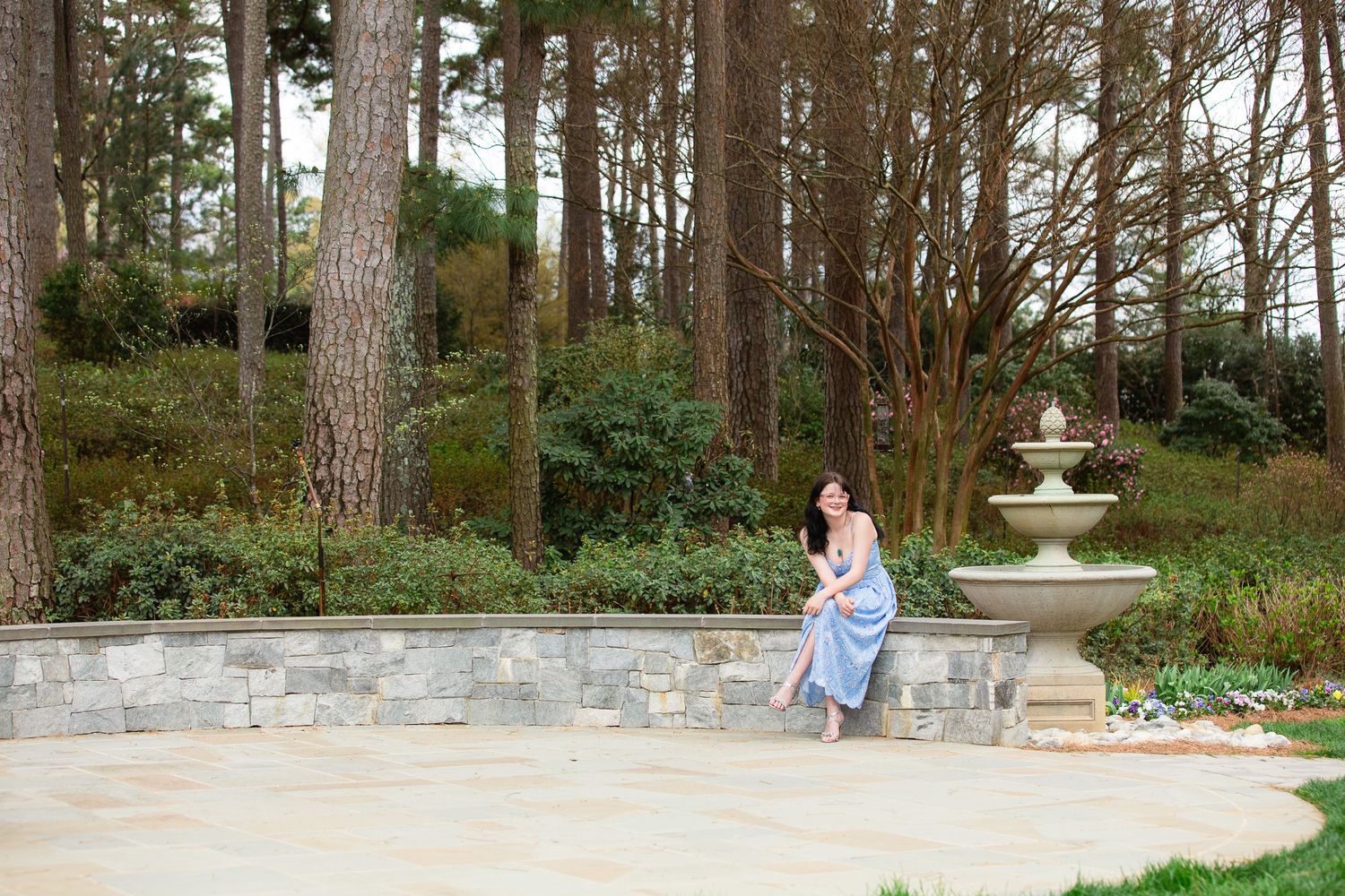 Woman in blue dress sitting on stone wall by fountain surrounded by tall pine trees in a serene garden setting for senior photos