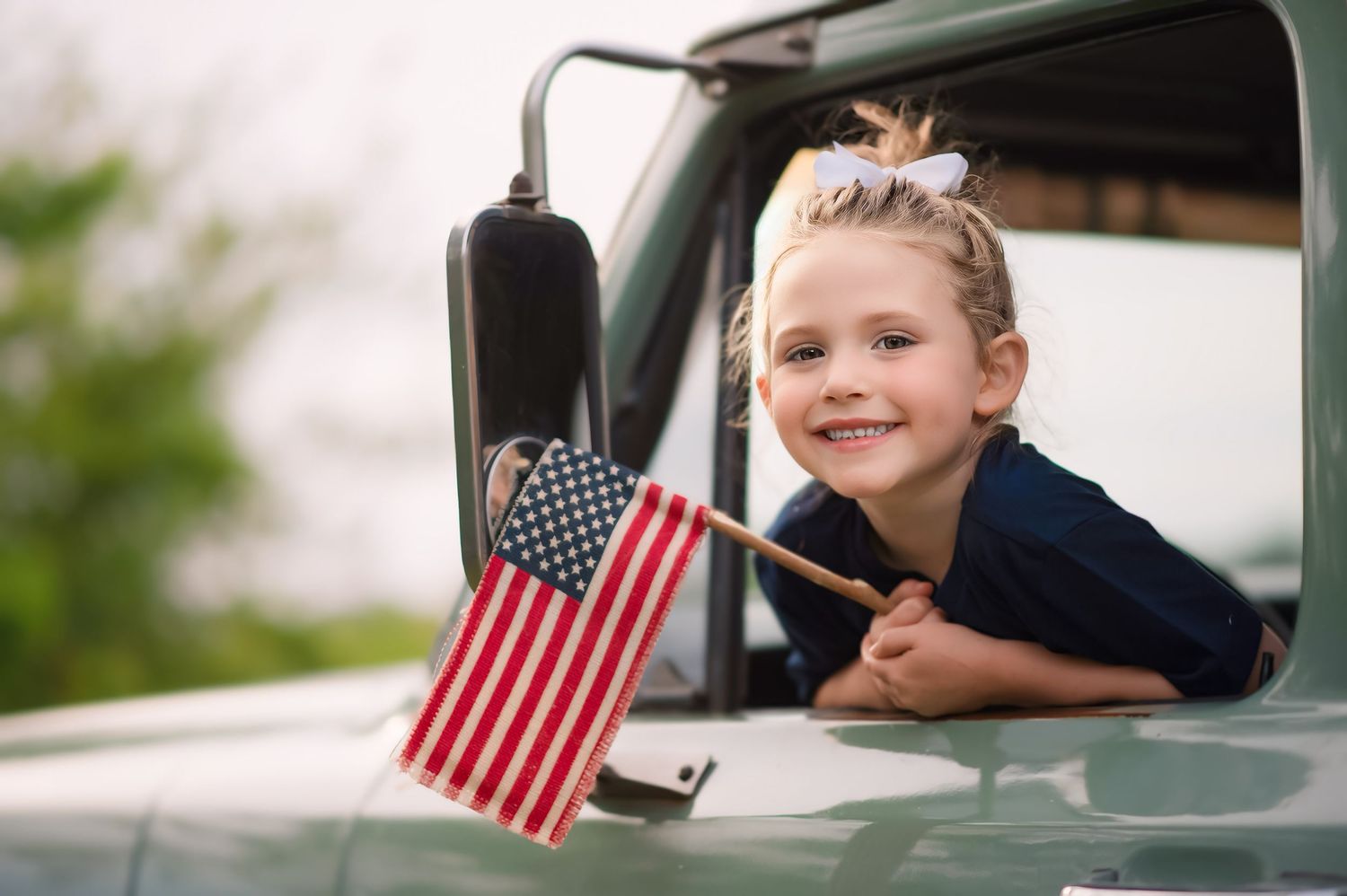 Young girl smiling and holding an American flag while leaning out of a vintage green truck window.