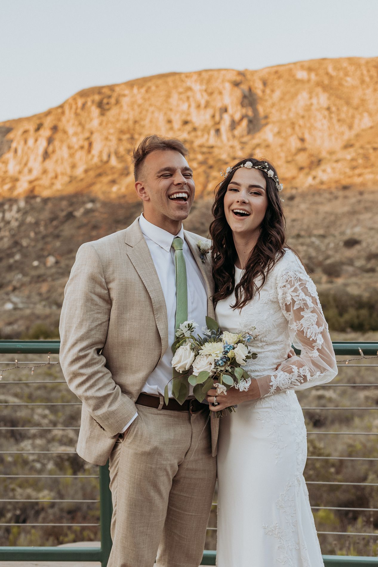 Smiling bride and groom in beige suit with green tie holding white bouquet, laughing outdoors with mountain backdrop at Mission Trails.