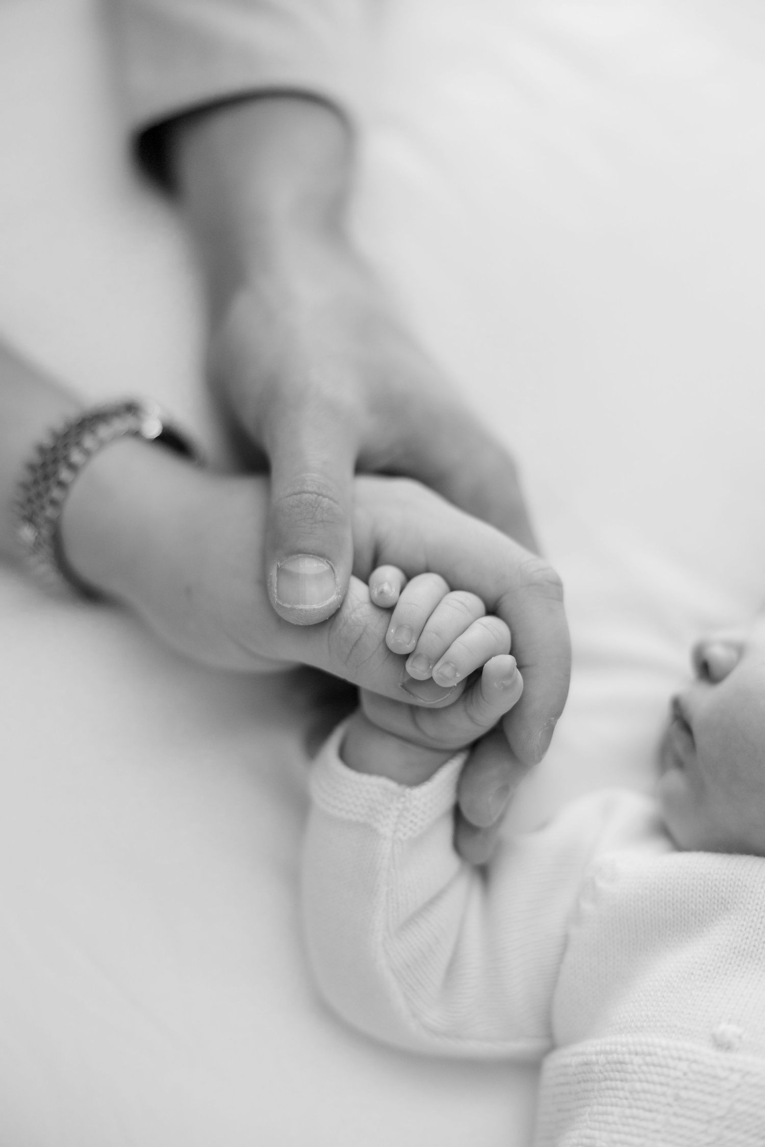 Black and white photo of a newborn baby holding an adult finger, with a bracelet visible on the parent's wrist.