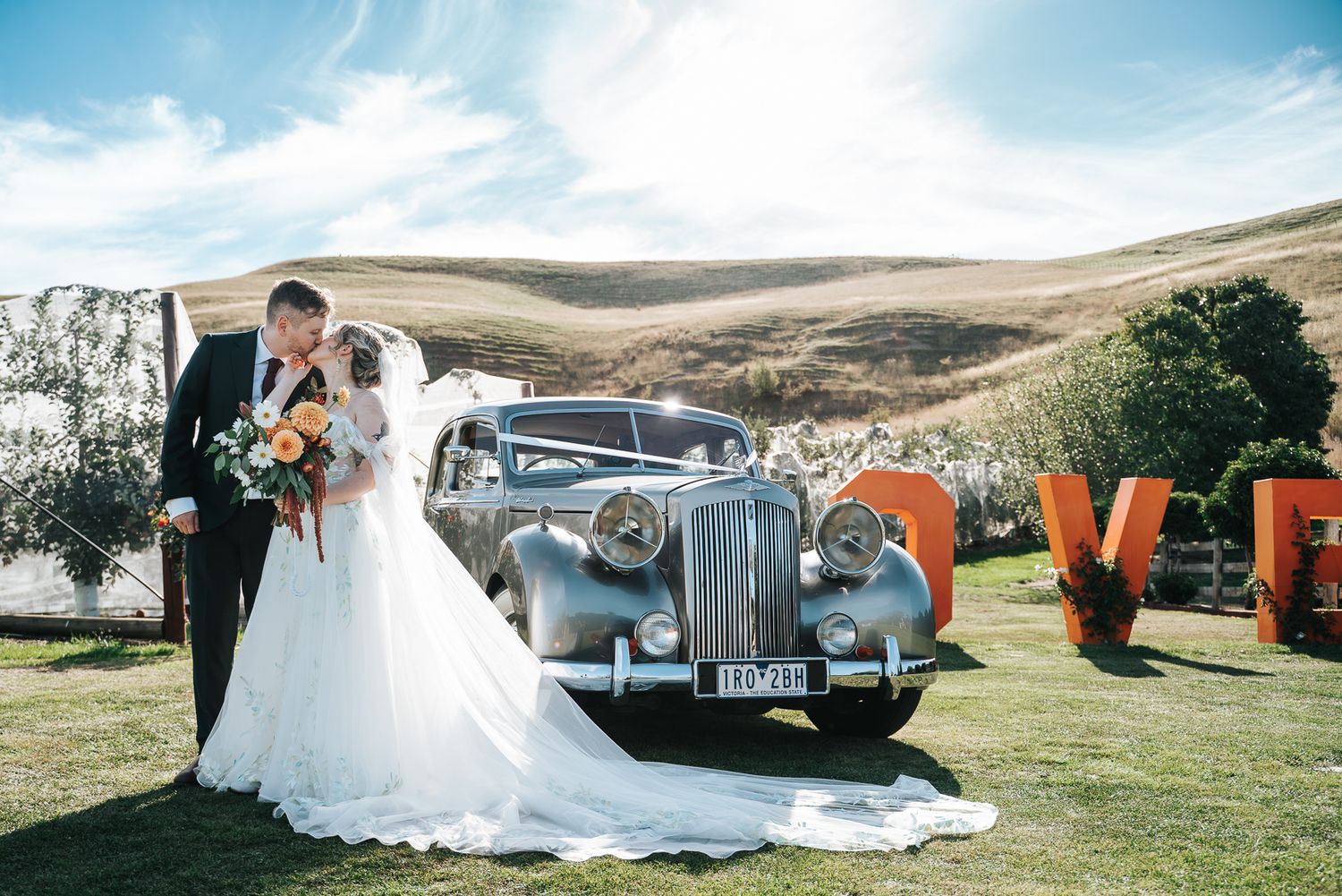 Bride and groom pose beside a vintage silver car with LOVE letters on their outdoor wedding day taken by a Melbourne Wedding Photographer.