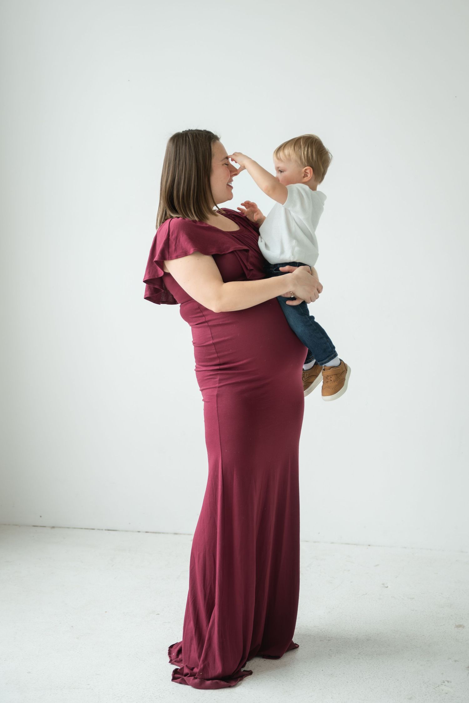 Pregnant woman in burgundy gown holds toddler boy in white shirt, sharing a tender moment in a bright studio.