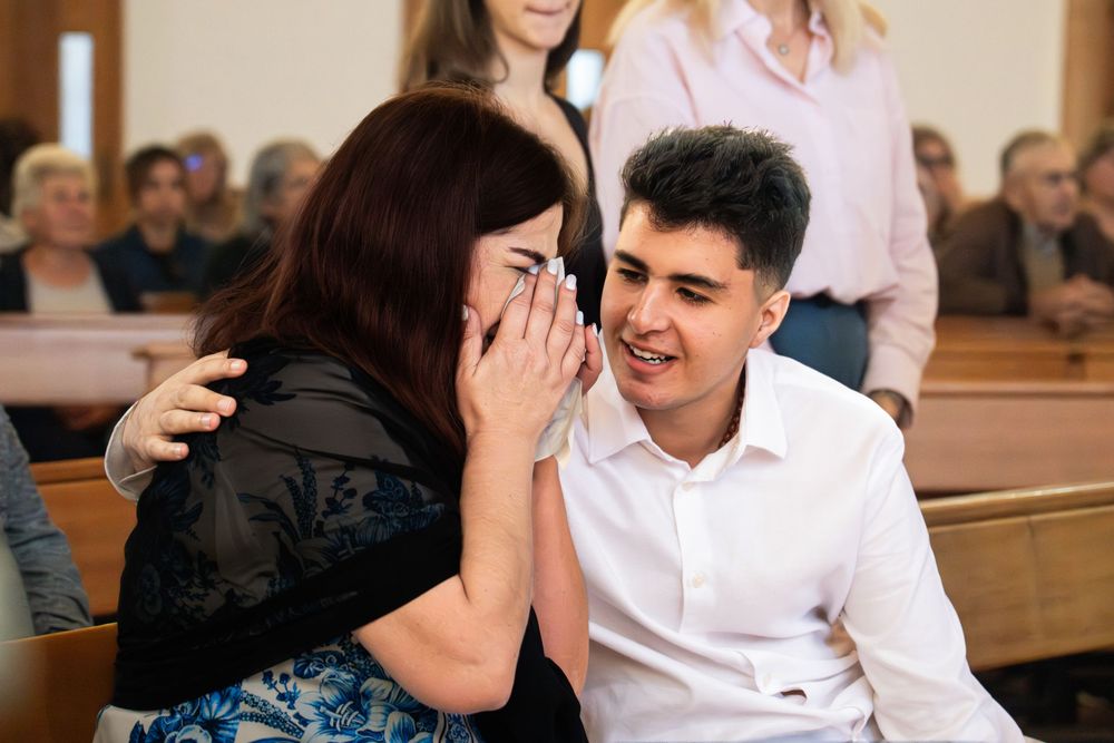 Woman whispering to smiling young man in white shirt while seated on church pew during a ceremony.