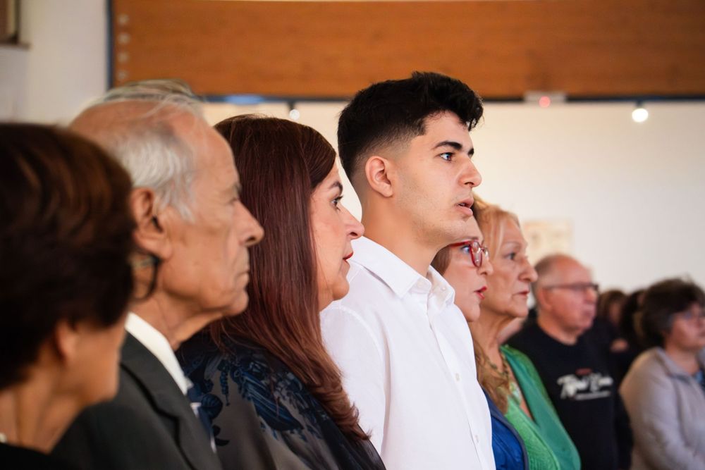 A diverse group of people standing in profile view during a formal ceremony inside a bright community hall.