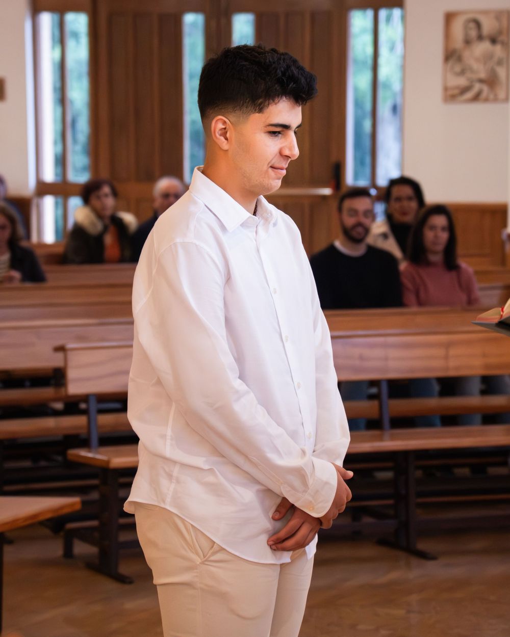 Young man in white shirt and pants standing in a church during a religious ceremony with congregation seated behind him.