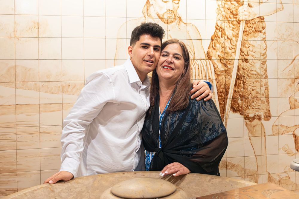 Two people smiling and posing together in front of an ornate tiled wall with artistic engravings.