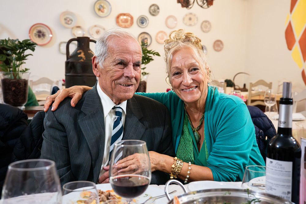 Elderly couple smiling together at a restaurant dinner table with wine glasses and a bottle of red wine.