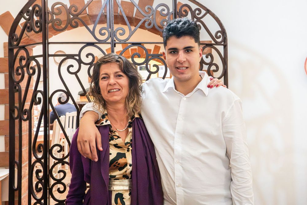 A woman and young man smile together in front of an ornate wrought iron gate at a formal celebration.