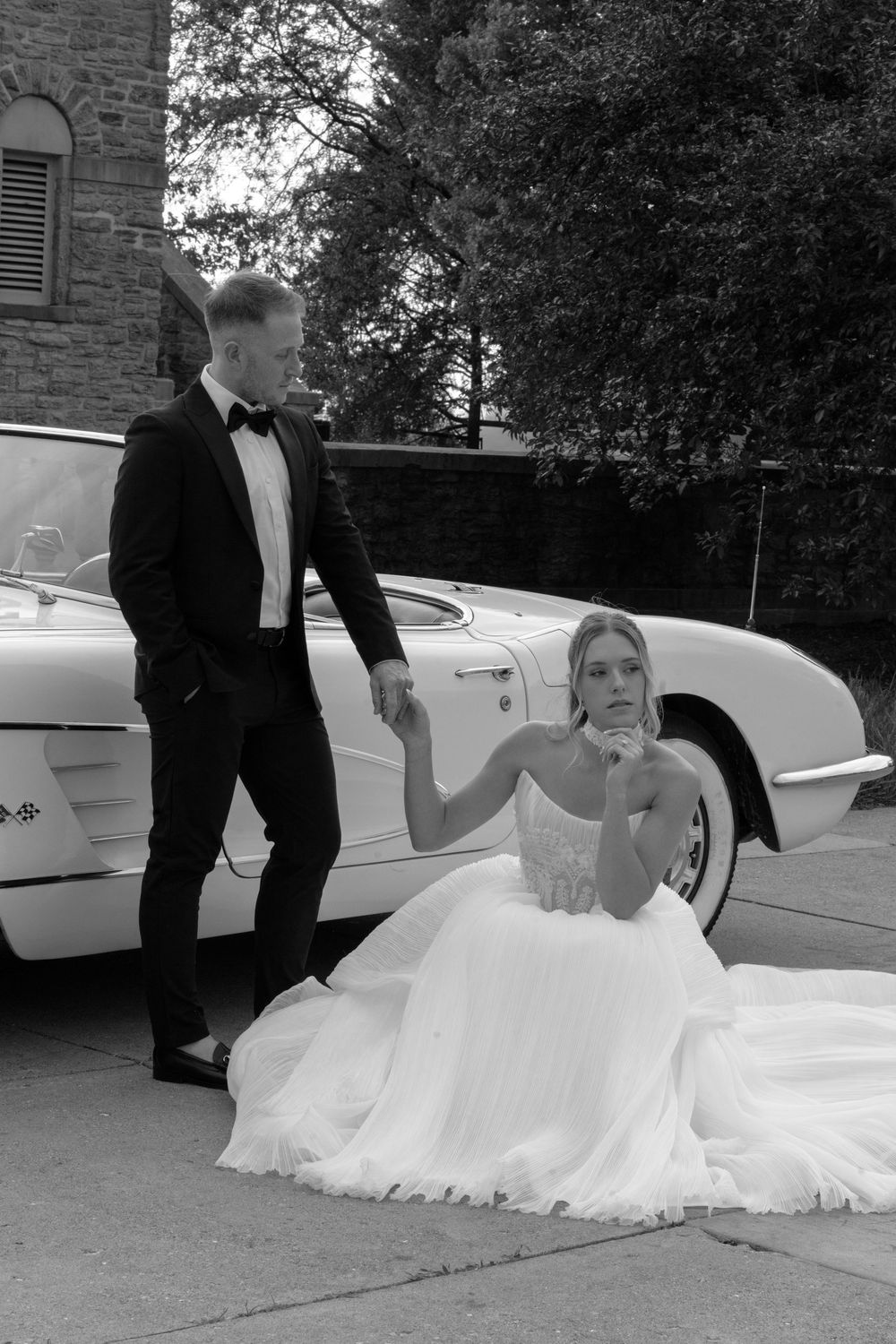 Bride in white gown sits beside vintage white Corvette as groom in tuxedo stands nearby in black and white photo.