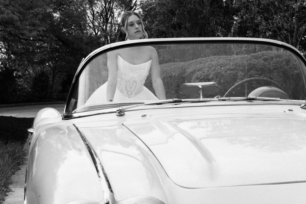 Elegant bride in white gown posing in a classic convertible car on a tree-lined road, black and white photo.